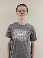 A young person with short hair and glasses stands against a plain light-colored wall, wearing a grey t-shirt with a geometric graphic on the front.