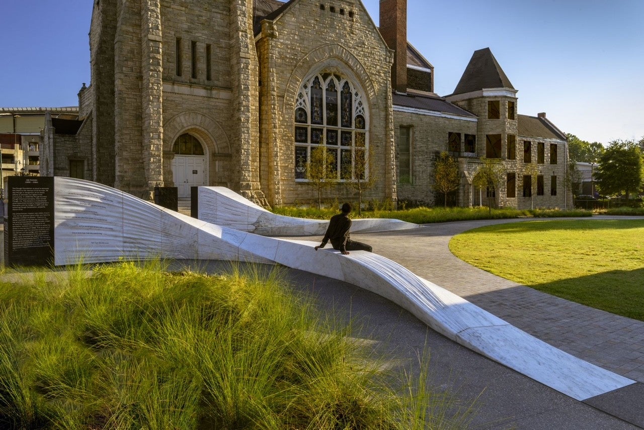 A person sits on a curving white marble bench in front of a large stone building with arched windows and a manicured lawn.