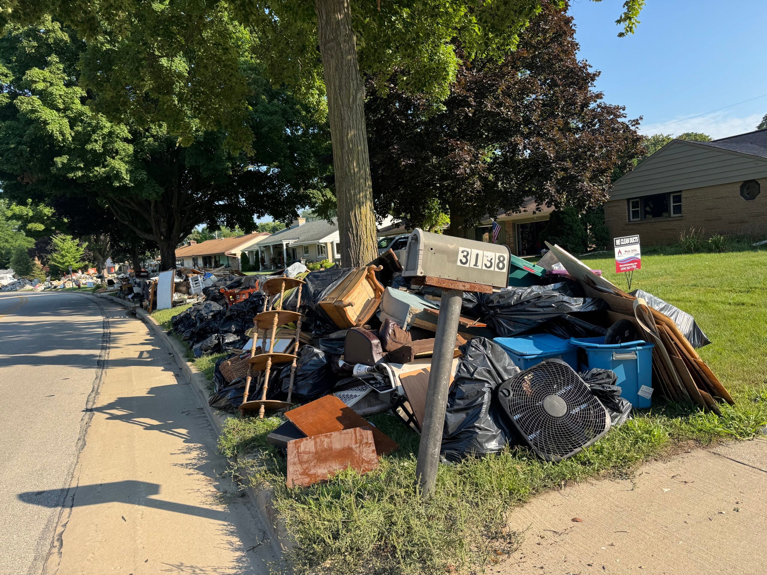Large pile of discarded household items and garbage bags sits curbside near a mailbox labeled 3138, in front of suburban houses on a sunny day.