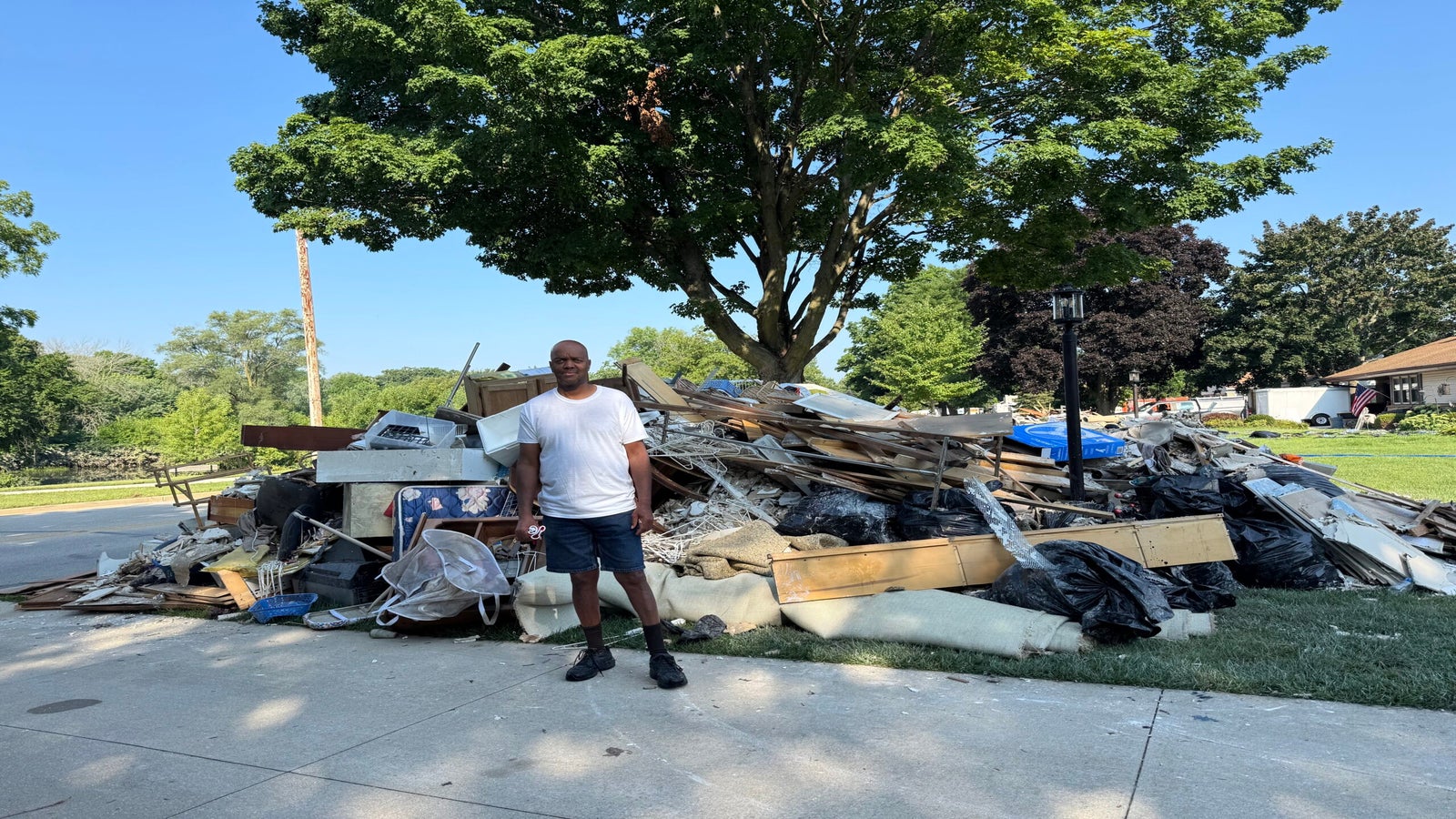 A man stands on a sidewalk in front of a large pile of debris and damaged household items, with trees and houses visible in the background.