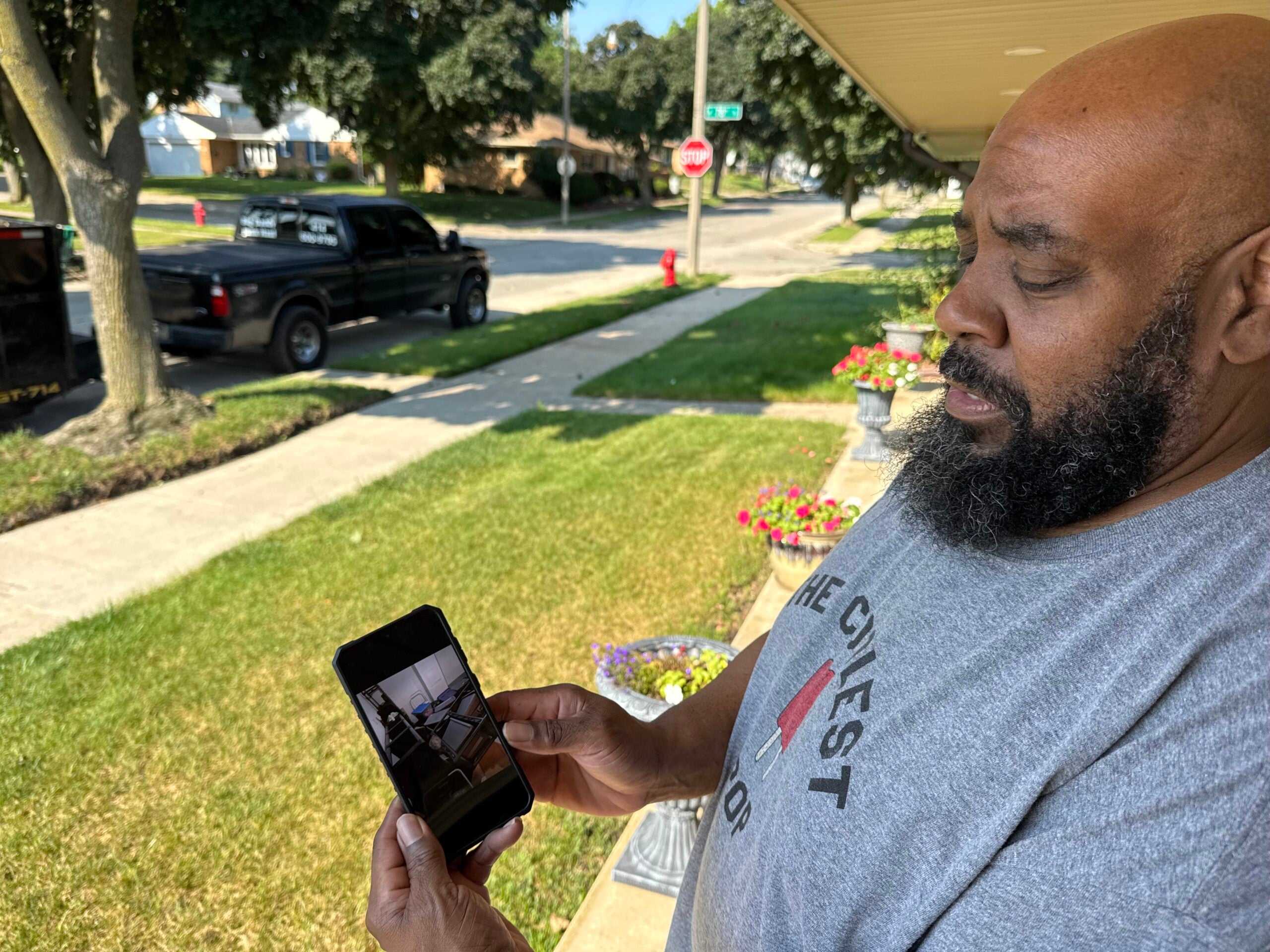 A man standing on a lawn looks at a photo on his smartphone, with a residential street, black pickup truck, and houses visible in the background.
