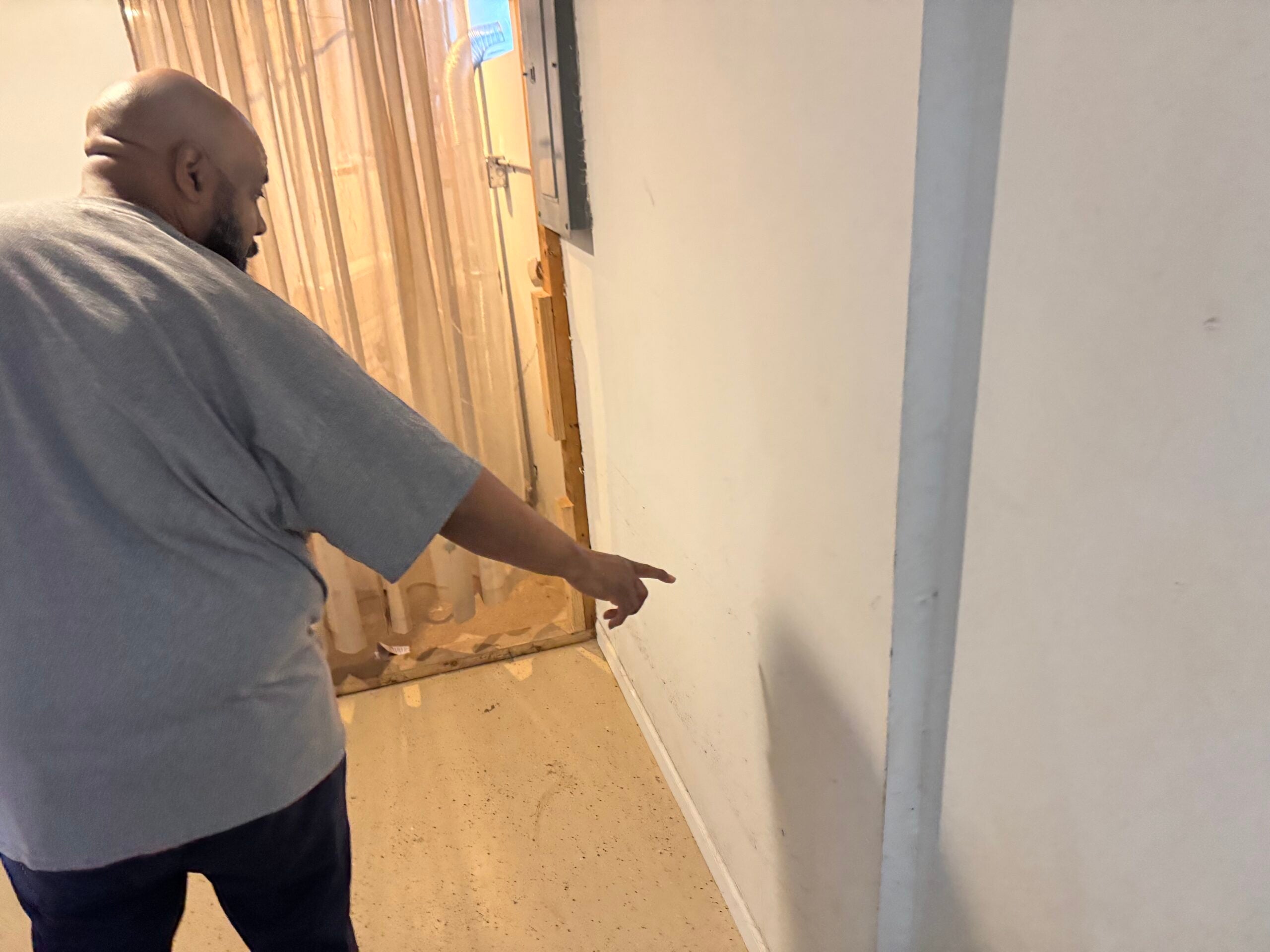 A person in a gray shirt points toward a blank white wall in a hallway with a beige curtain in the background.