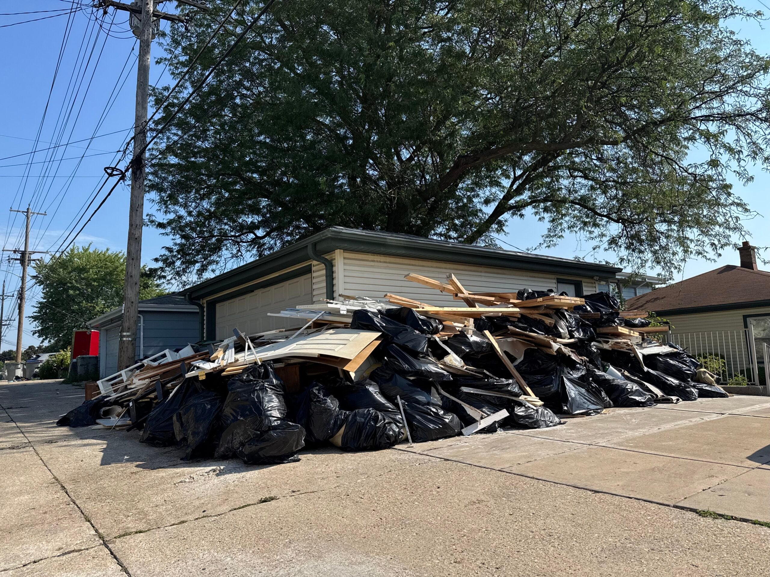 A large pile of black garbage bags and debris sits in front of a garage in an alley, with a tree and power lines in the background.