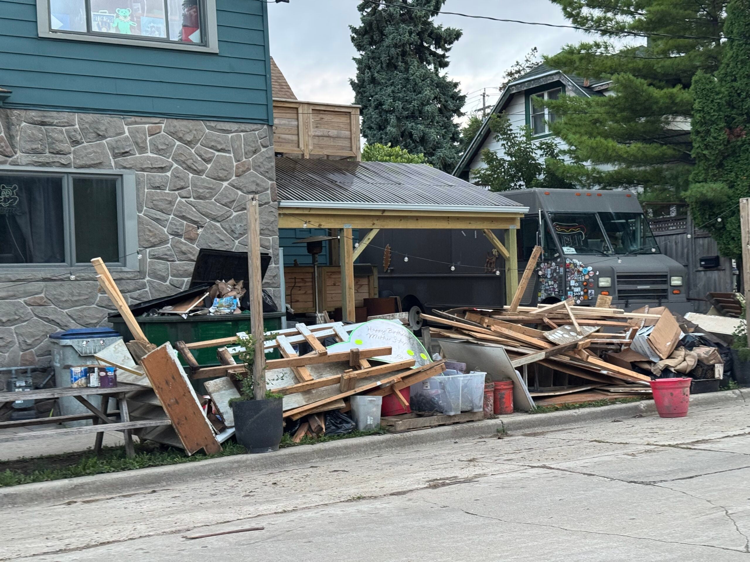A pile of wood debris, discarded furniture, and trash bins are stacked in front of a house with a covered porch near a delivery truck on a residential street.