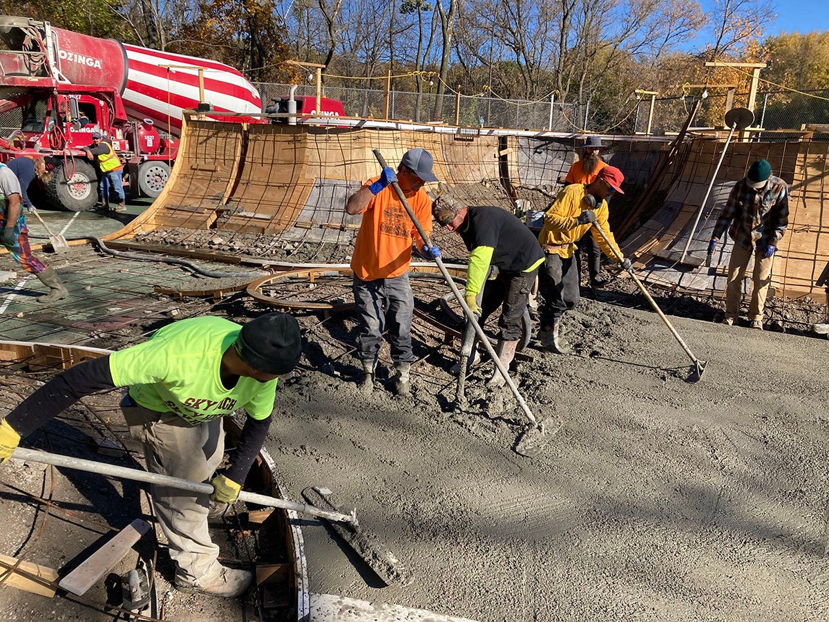 Five construction workers in safety gear are spreading and leveling wet concrete with tools at an outdoor construction site with curved wooden forms.