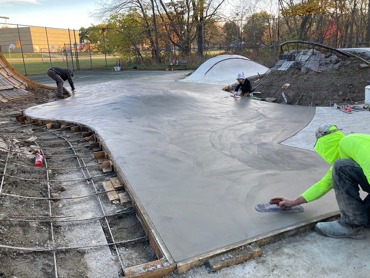 Workers smooth freshly poured concrete on a skatepark structure, with tools and materials visible around the construction area.