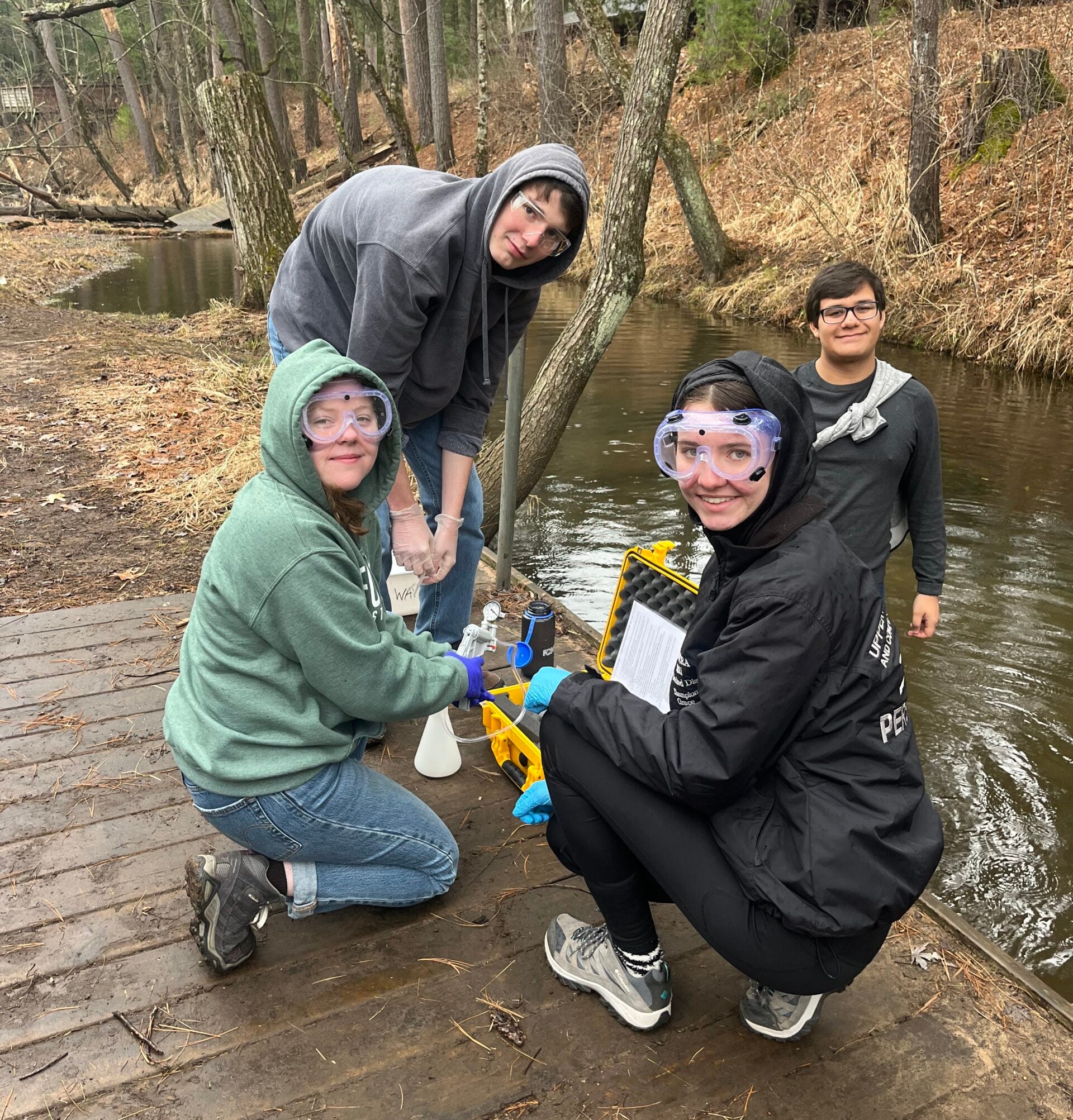 Four people wearing goggles and gloves collect water samples by a wooded creek, using scientific equipment on a wooden dock.