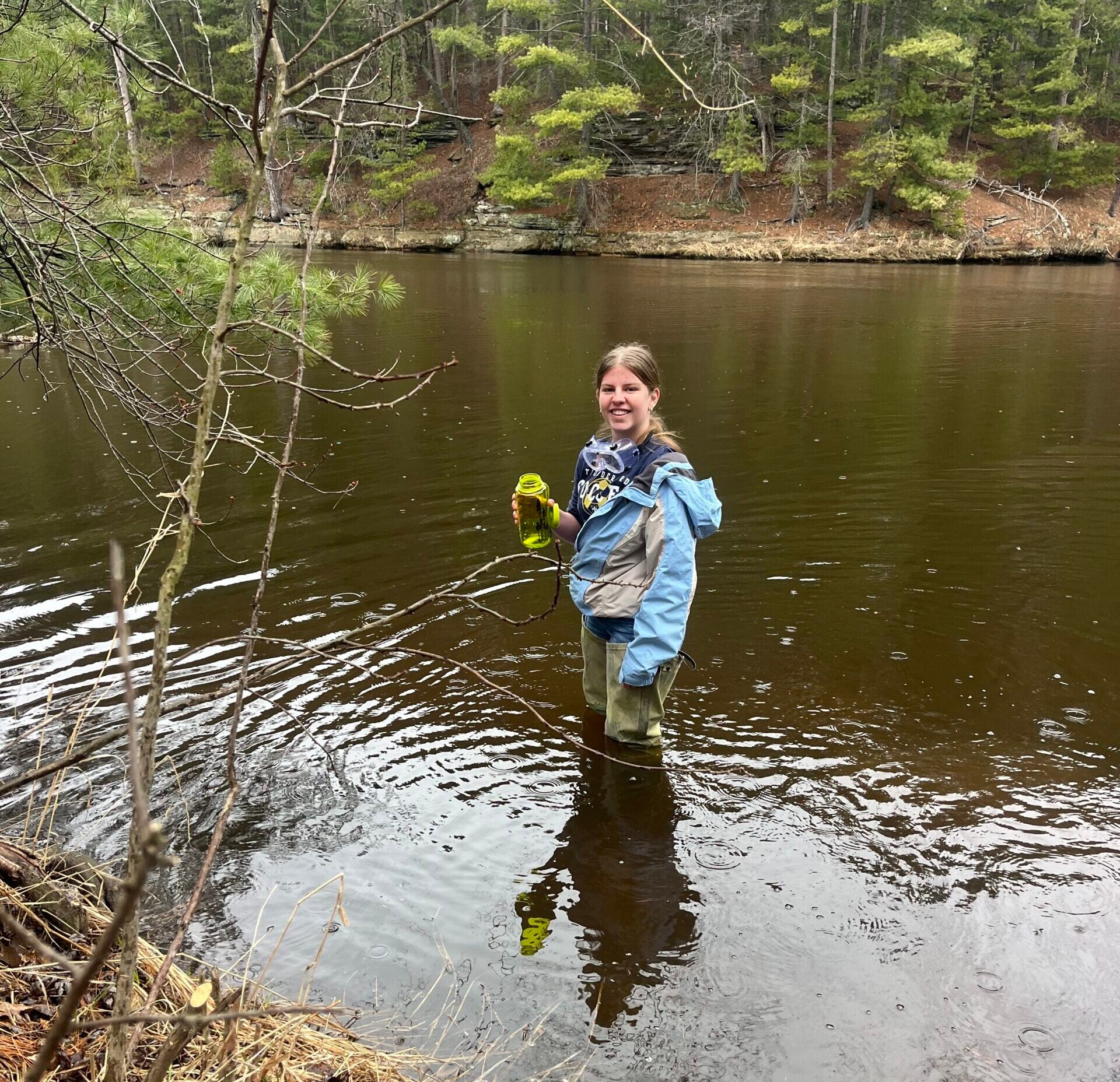 A person wearing waders stands knee-deep in a river, holding a yellow object, with a forested area in the background.
