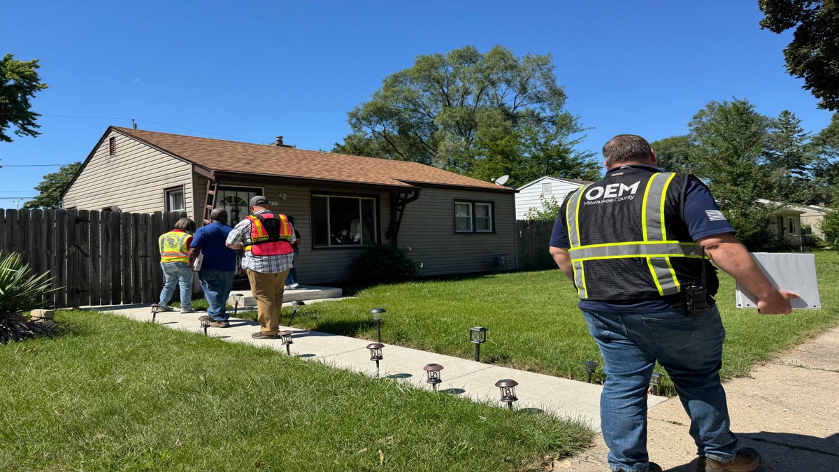 Four people wearing safety vests and carrying equipment walk toward the entrance of a small house on a sunny day.