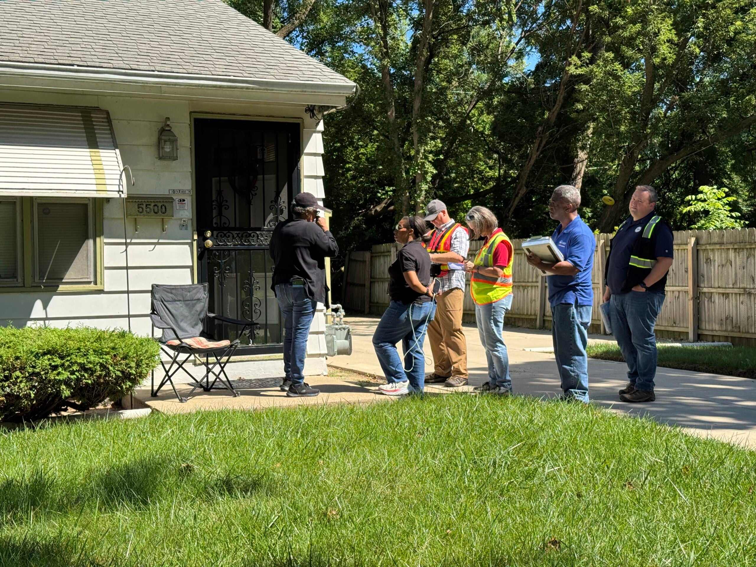 Six people, some in safety vests, stand outside a house near the front door, appearing to conduct an inspection or assessment on a sunny day.