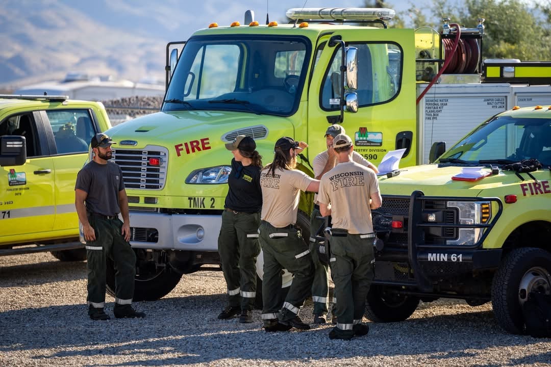 A group of firefighters in uniform stand and talk beside yellow fire trucks in a gravel lot under daylight.