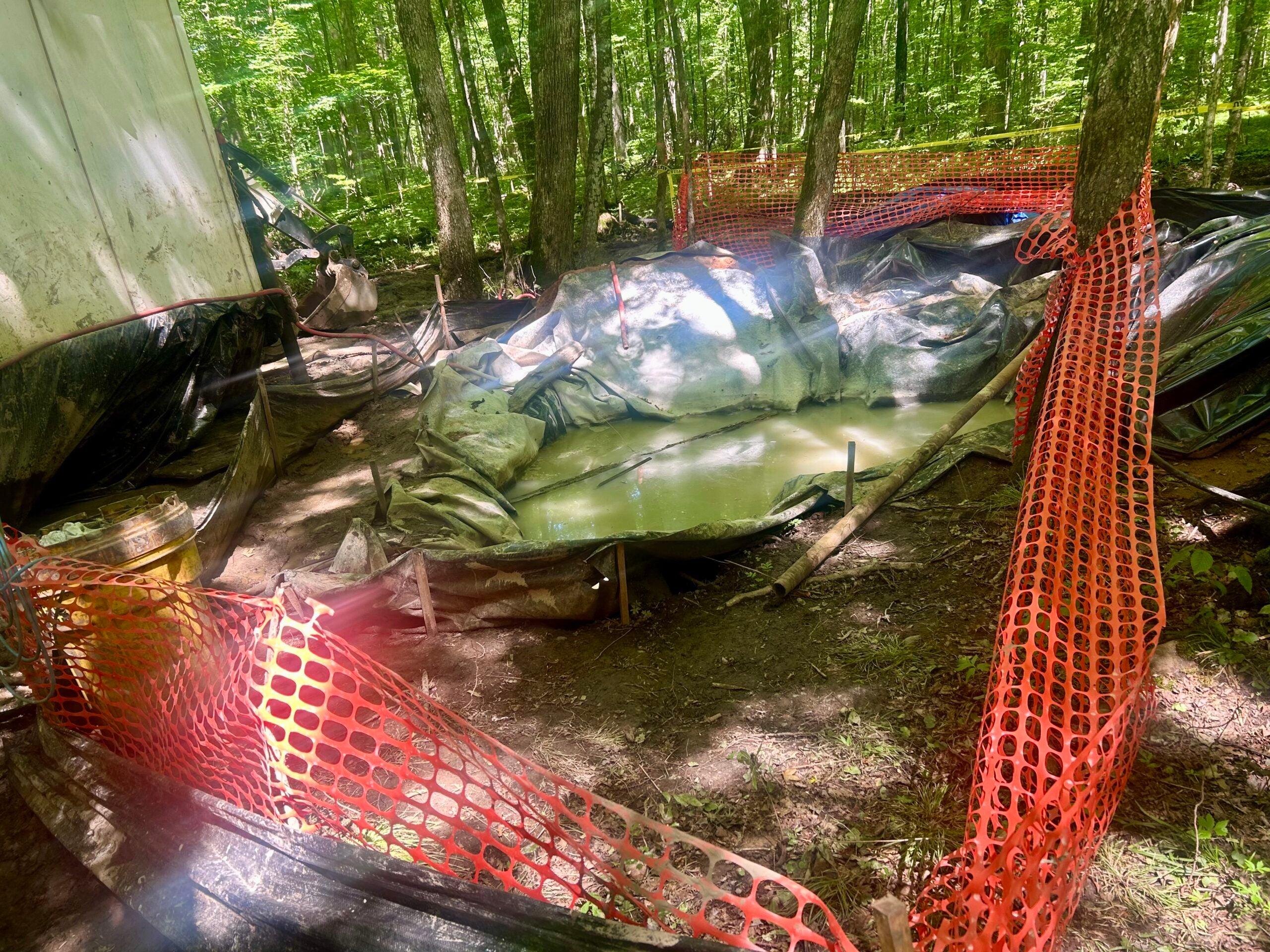 A muddy pit covered with tarps holds stagnant water, surrounded by orange safety fencing in a wooded area with sunlight filtering through trees.