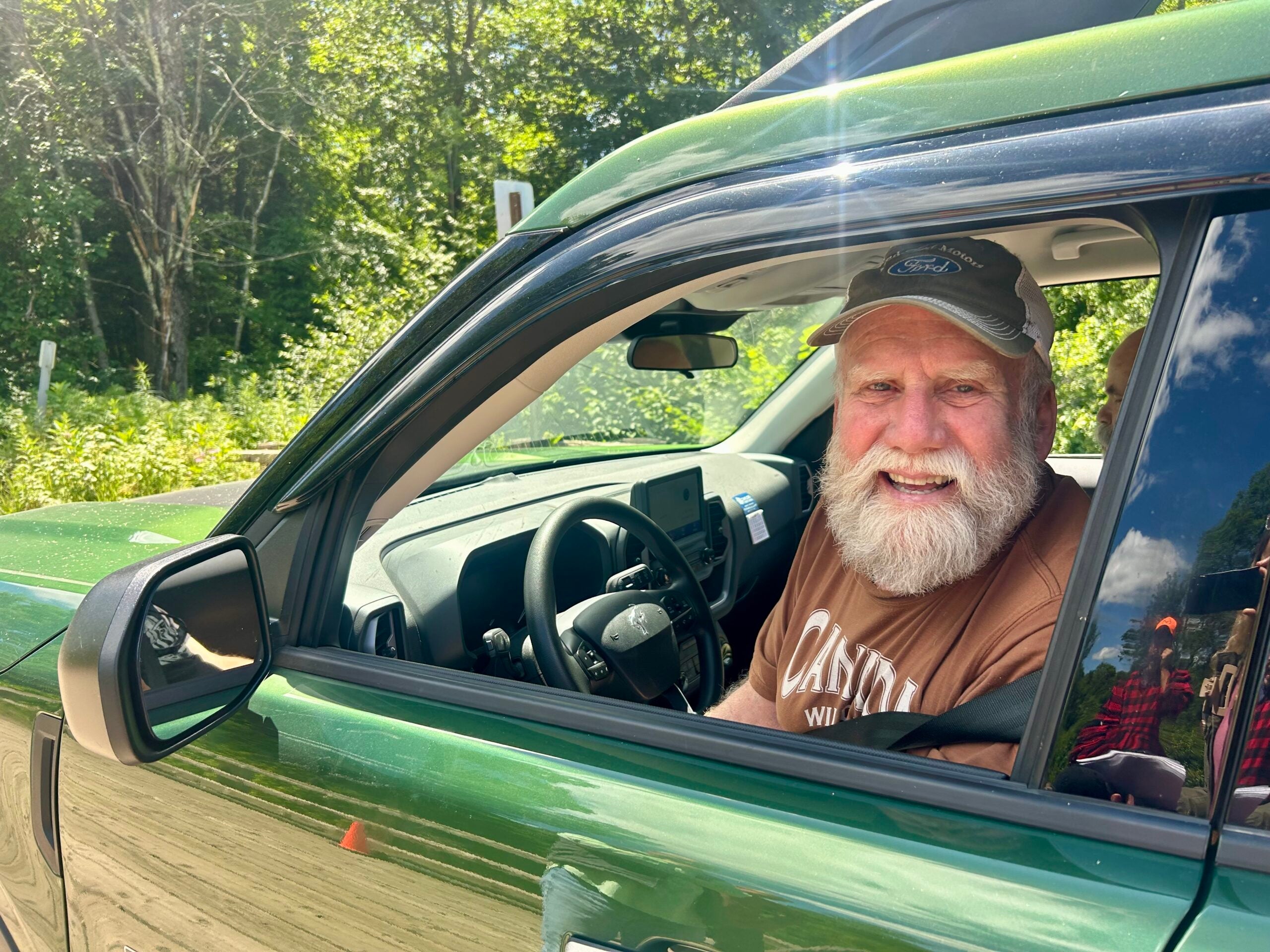 An older man with a white beard smiles while sitting in the driver’s seat of a green vehicle parked outdoors on a sunny day.