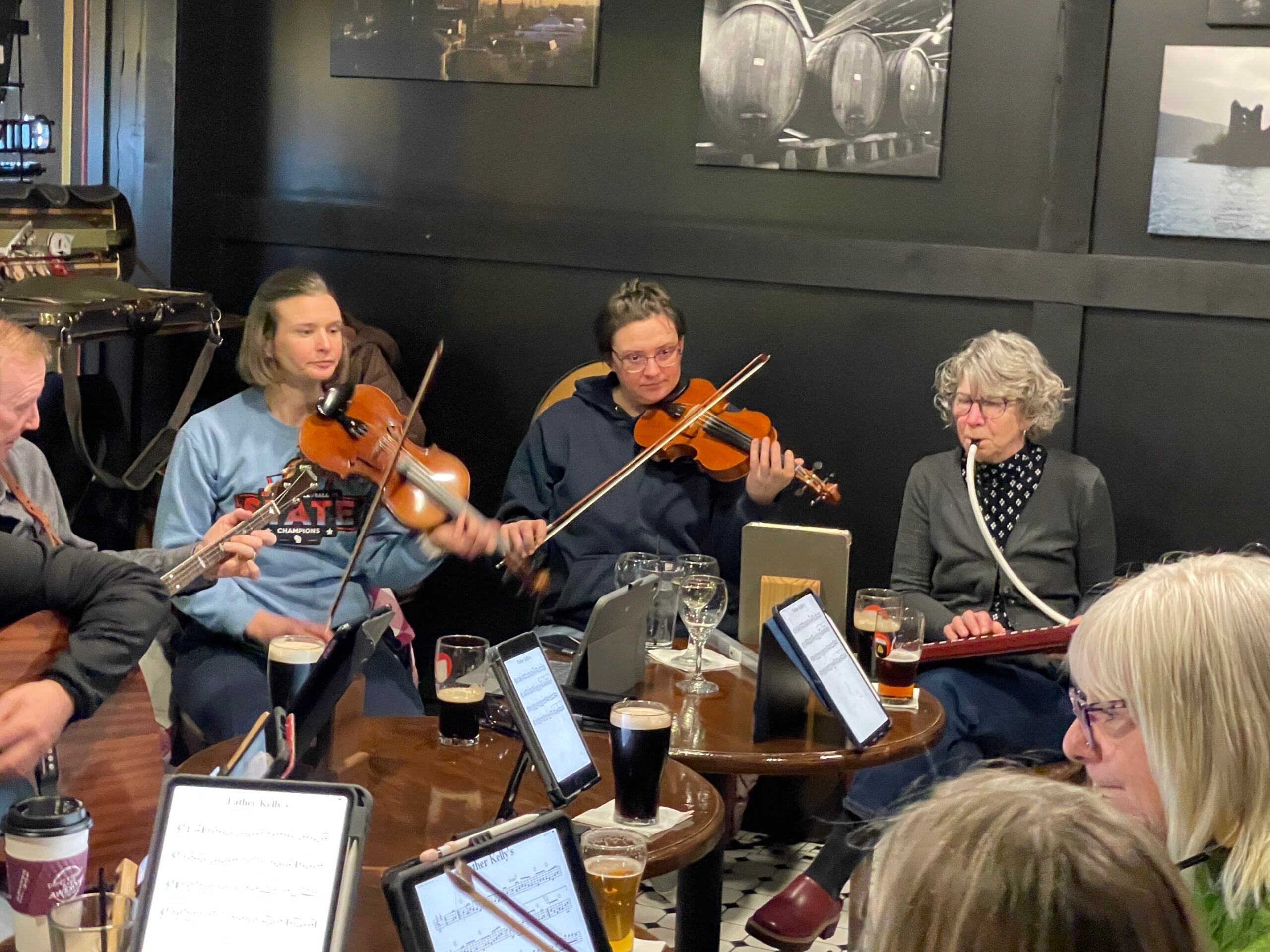 A group of people sit around a table in a pub, playing stringed instruments and reading sheet music, with drinks on the table.
