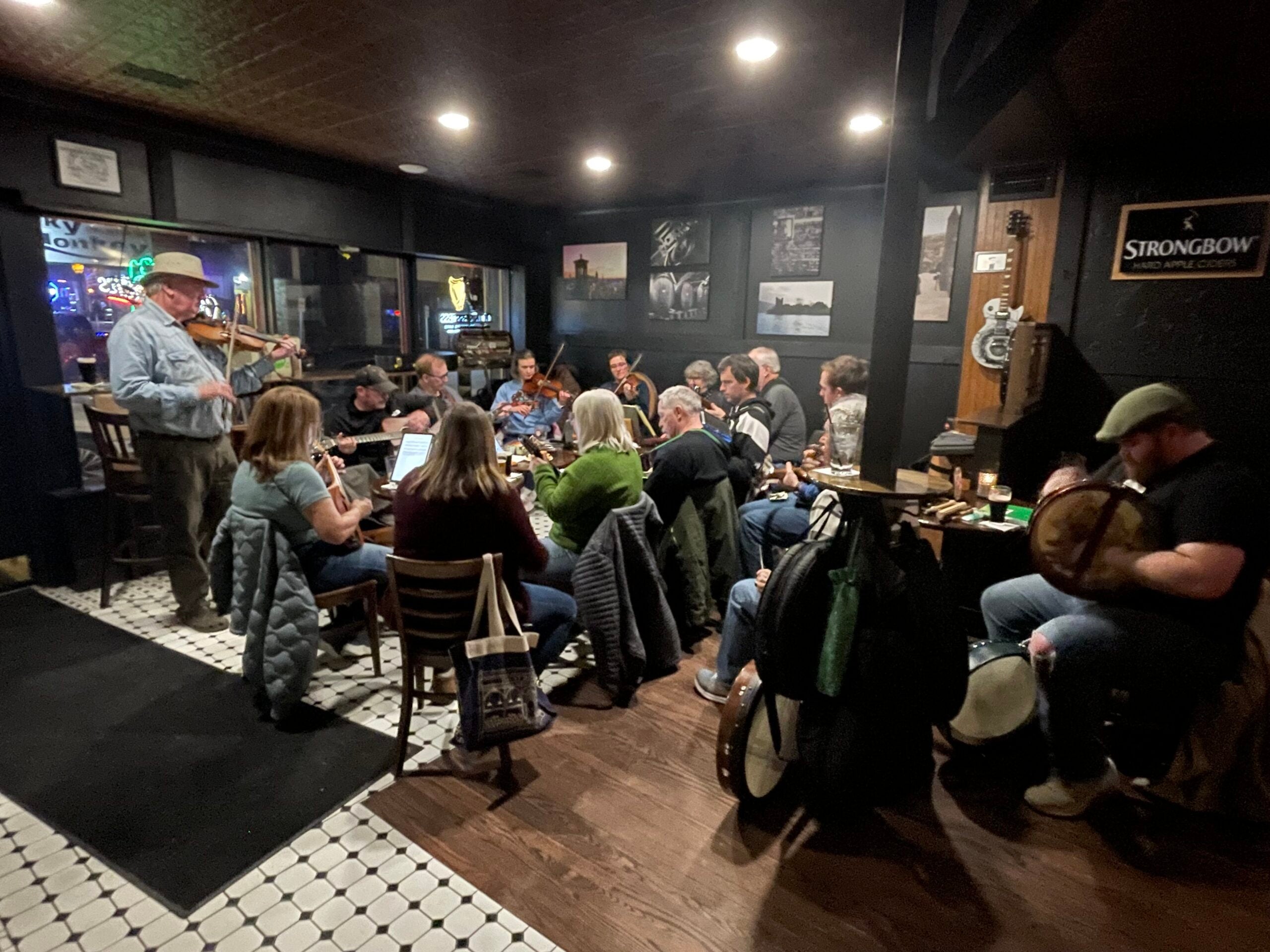 A group of people sit in a circle playing musical instruments in a dimly lit pub, with one man standing and playing the violin.