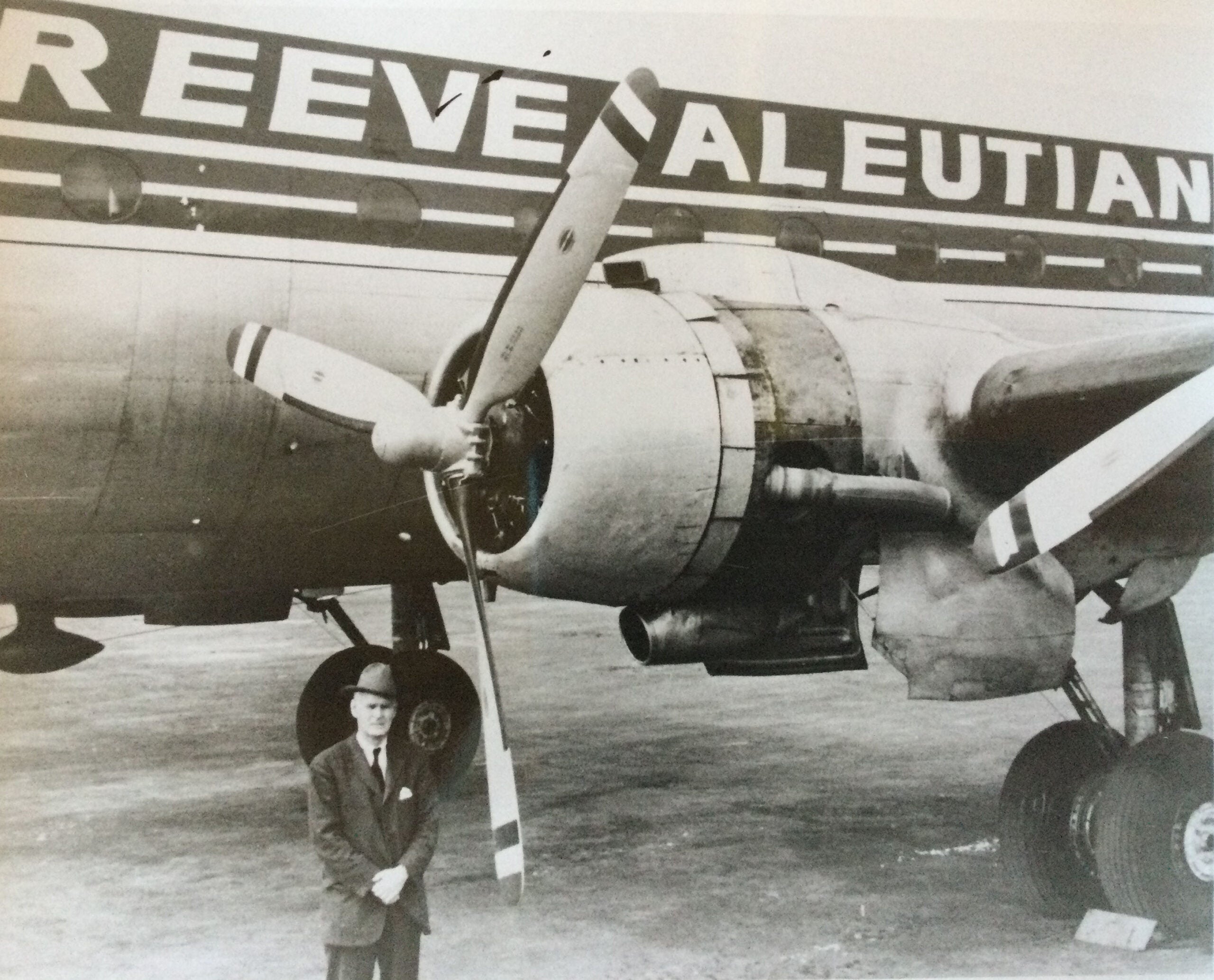 A man in a suit and hat stands in front of an airplane engine; the planes fuselage displays the text REEVE ALEUTIAN.