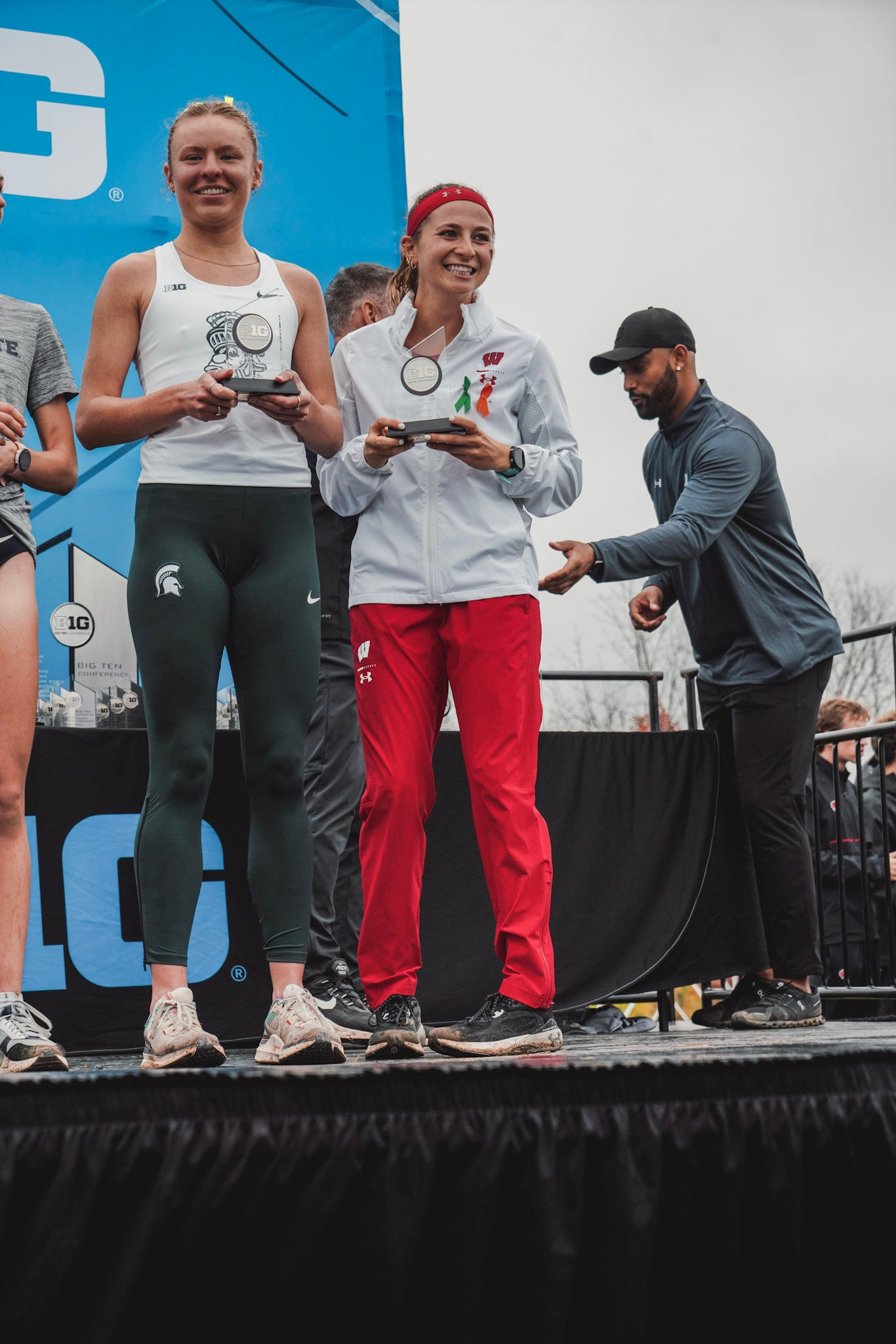Two female athletes stand on a podium holding trophies, smiling. A man in a cap stands nearby. A blue banner with BIG text is visible in the background.