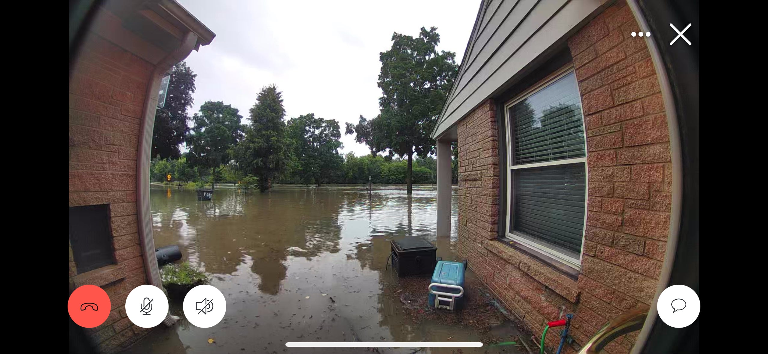 Floodwater surrounds a brick house and submerges the yard, reaching the base of the windows; trees and lawn equipment are partially underwater.