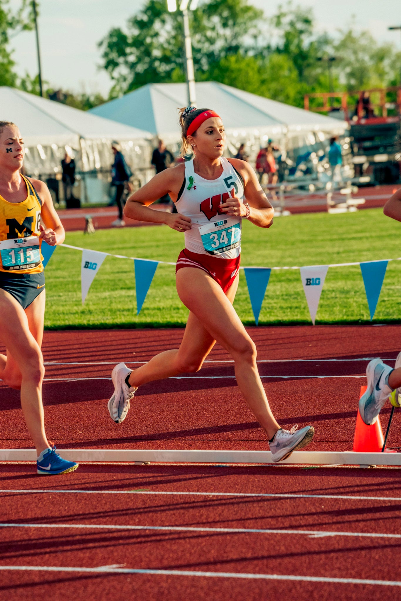 Three female athletes run on a track during a race, with one in a red and white uniform leading, and others in blue and yellow uniforms beside her. Tents and spectators are in the background.