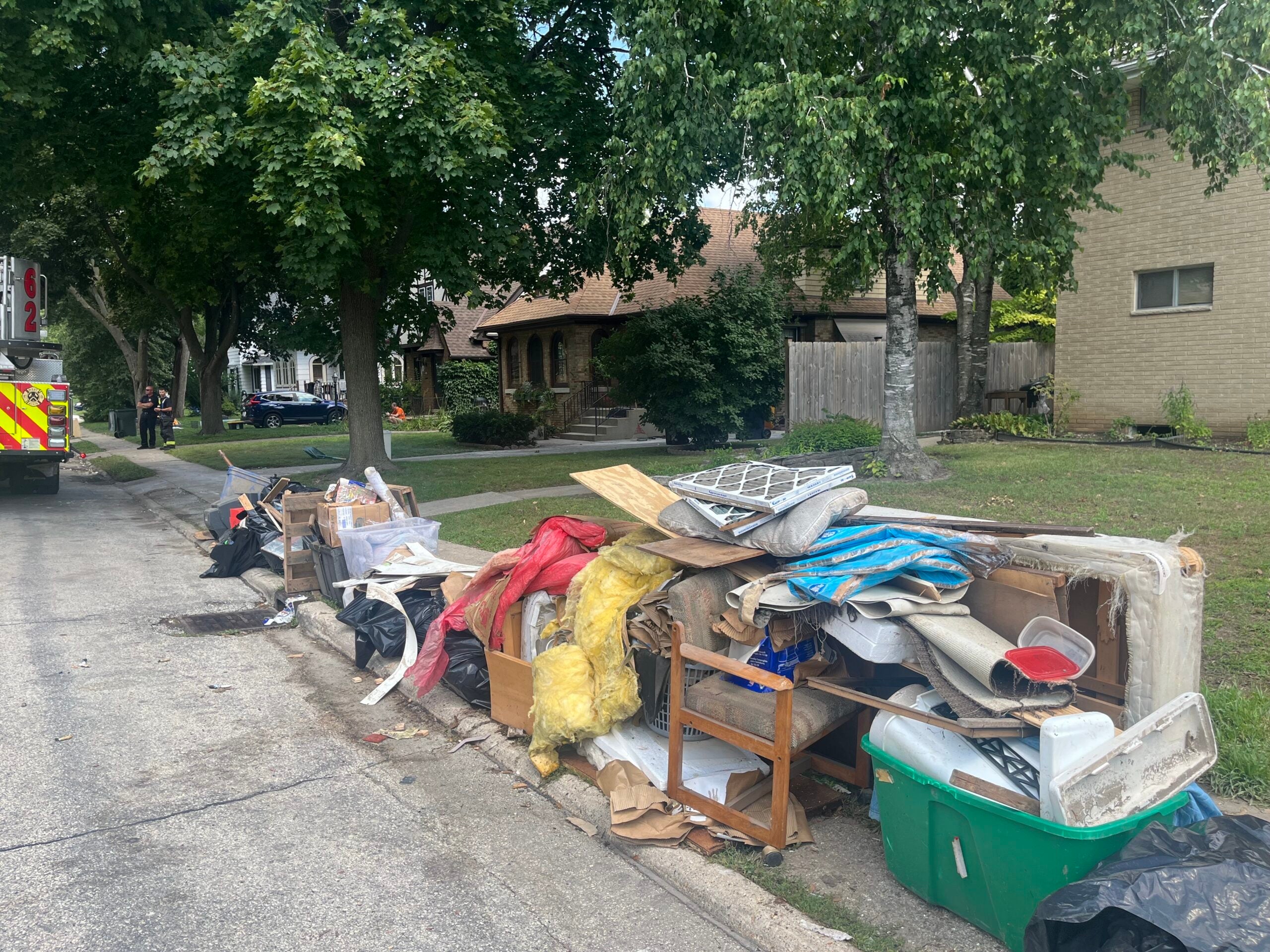 Large piles of assorted household debris, furniture, and trash line the curb of a residential street, with houses and trees in the background.