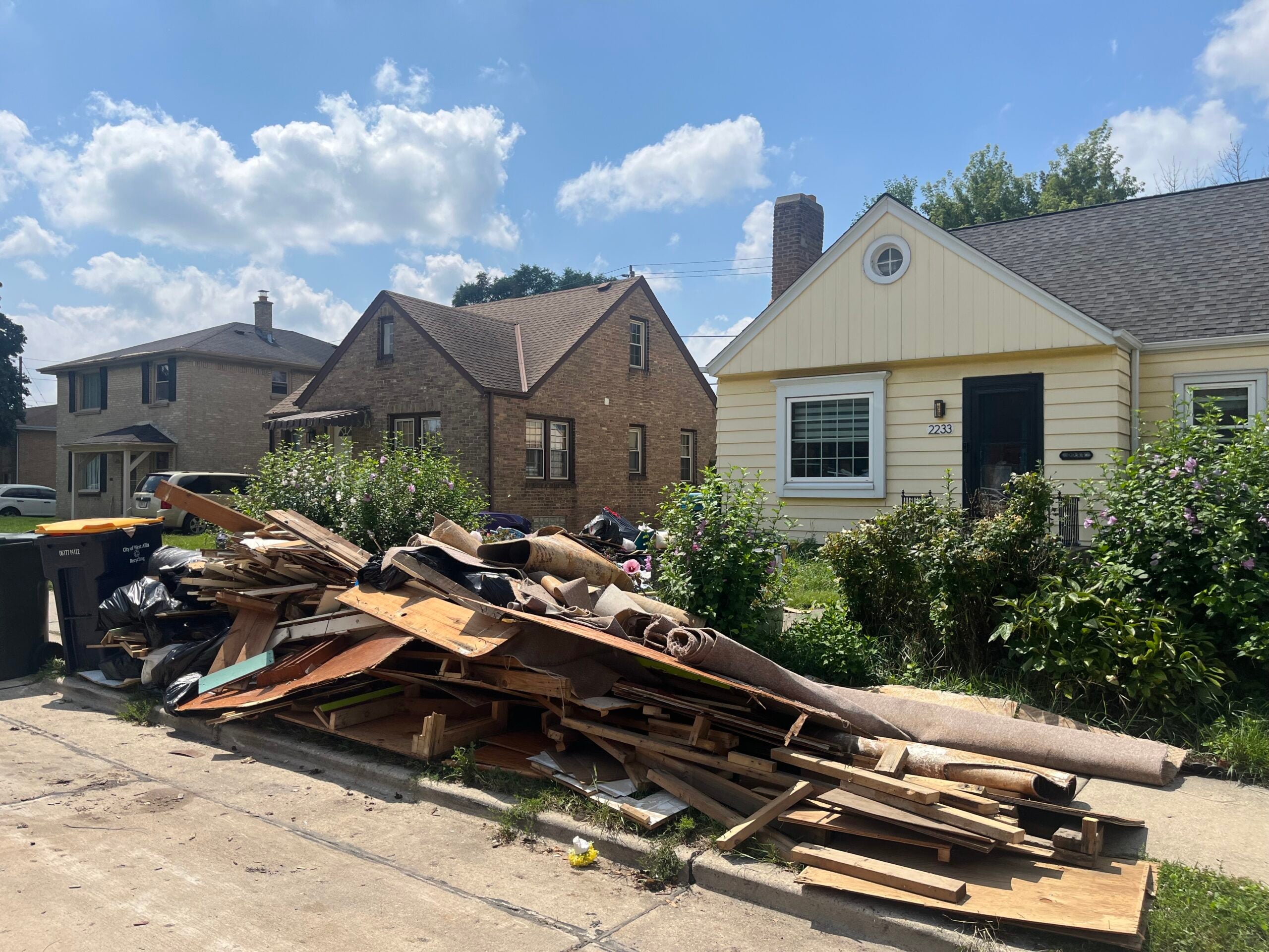 A large pile of discarded wood, carpet, and debris sits on the curb in front of a yellow house in a residential neighborhood.