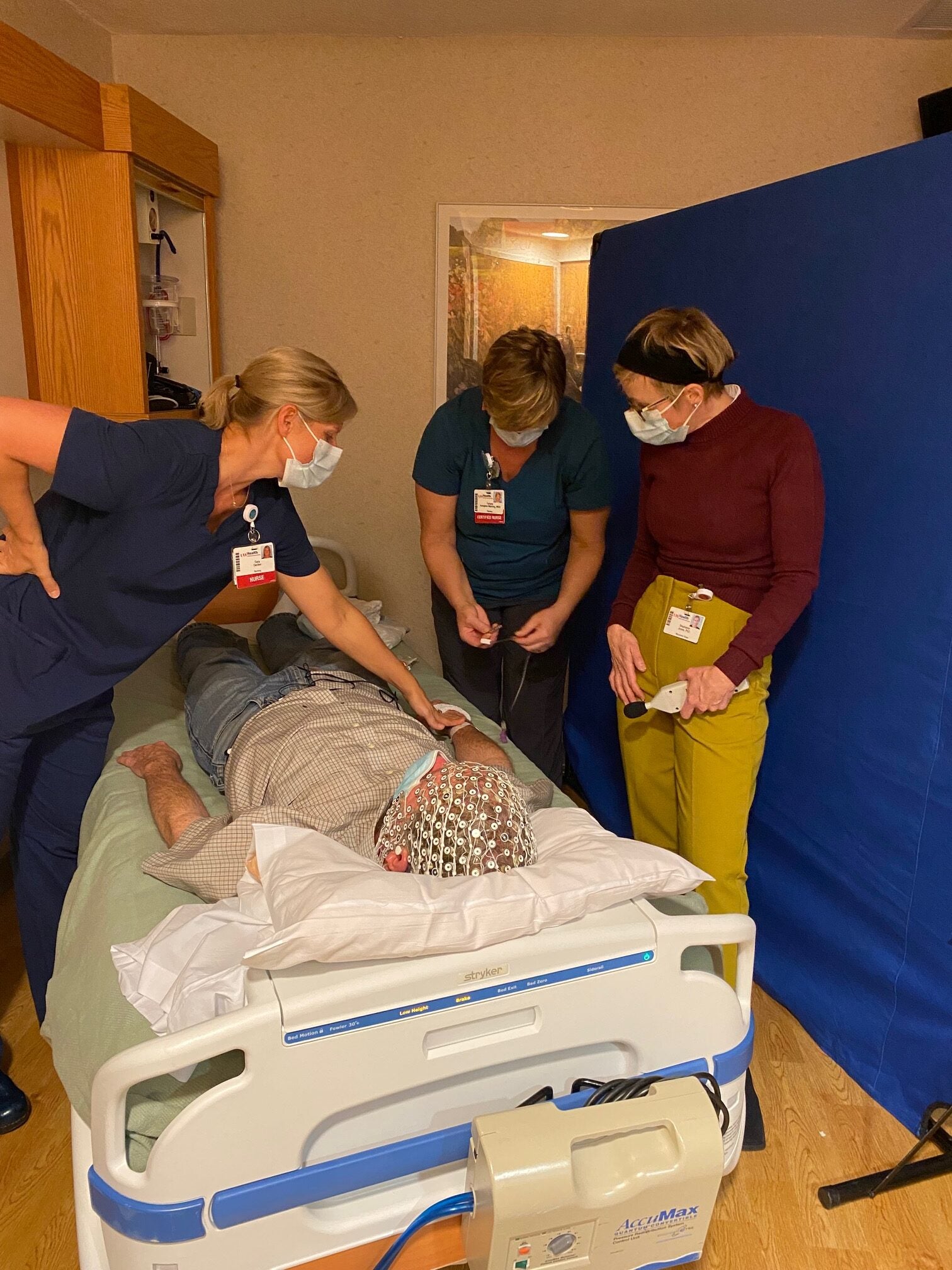 Three healthcare workers in masks stand around a patient lying face down on a hospital bed, monitoring him while he wears a medical cap with electrodes.