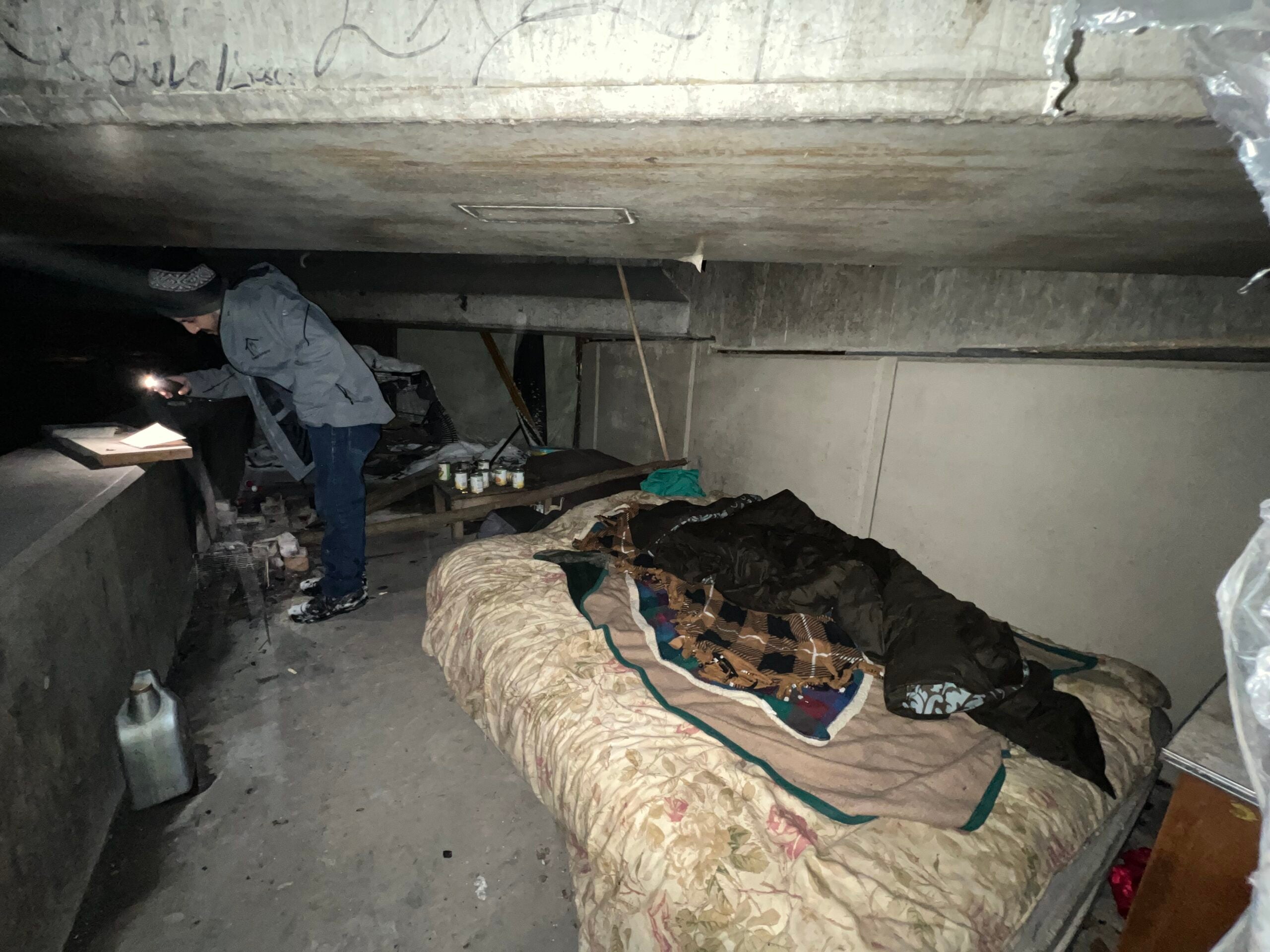 A person stands under a concrete overpass with a flashlight near a makeshift bed, blankets, and scattered belongings. The area appears to be used as a shelter.