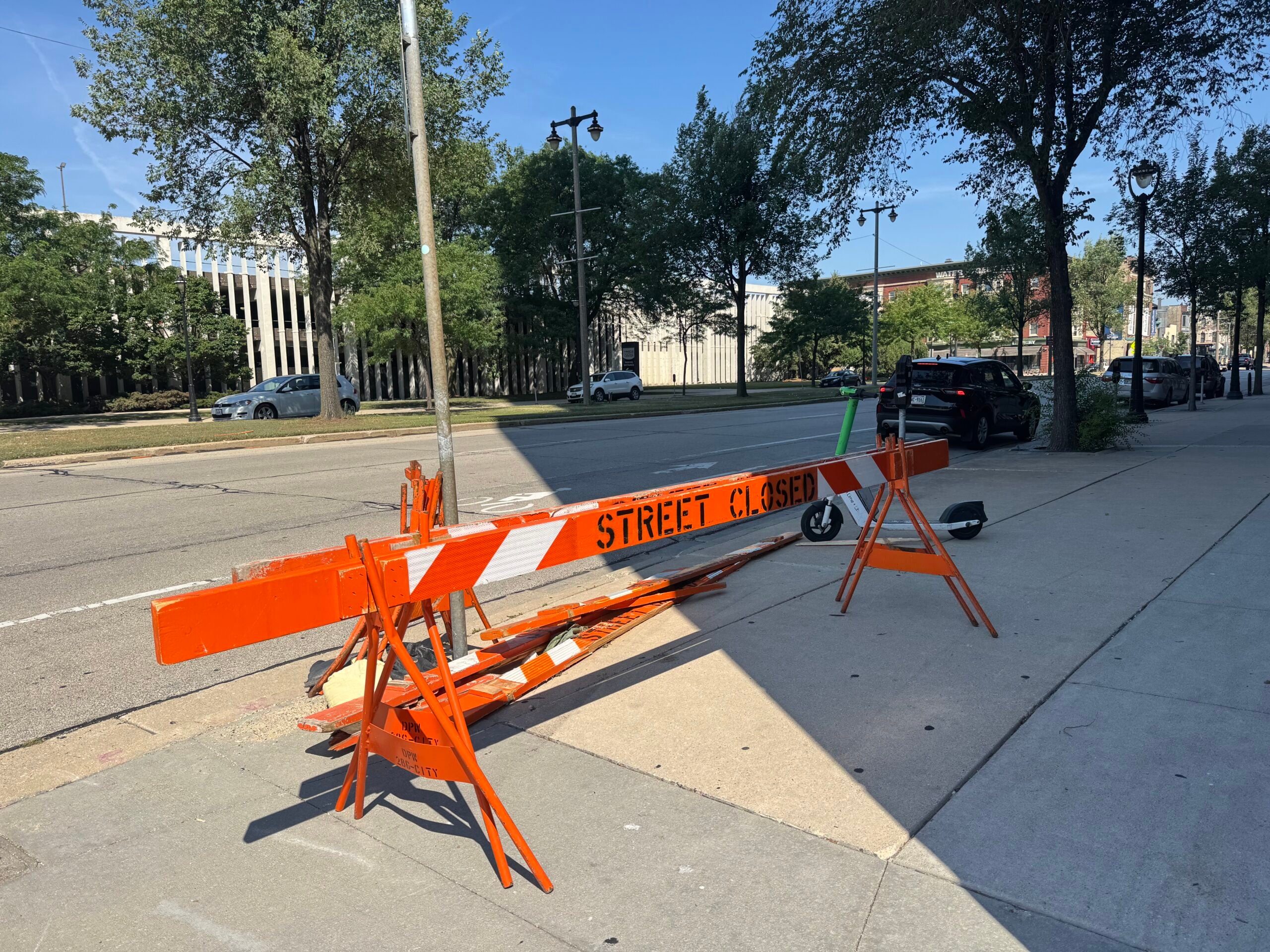 Damaged orange Street Closed barrier on sidewalk next to a road with trees and parked cars in the background on a sunny day.
