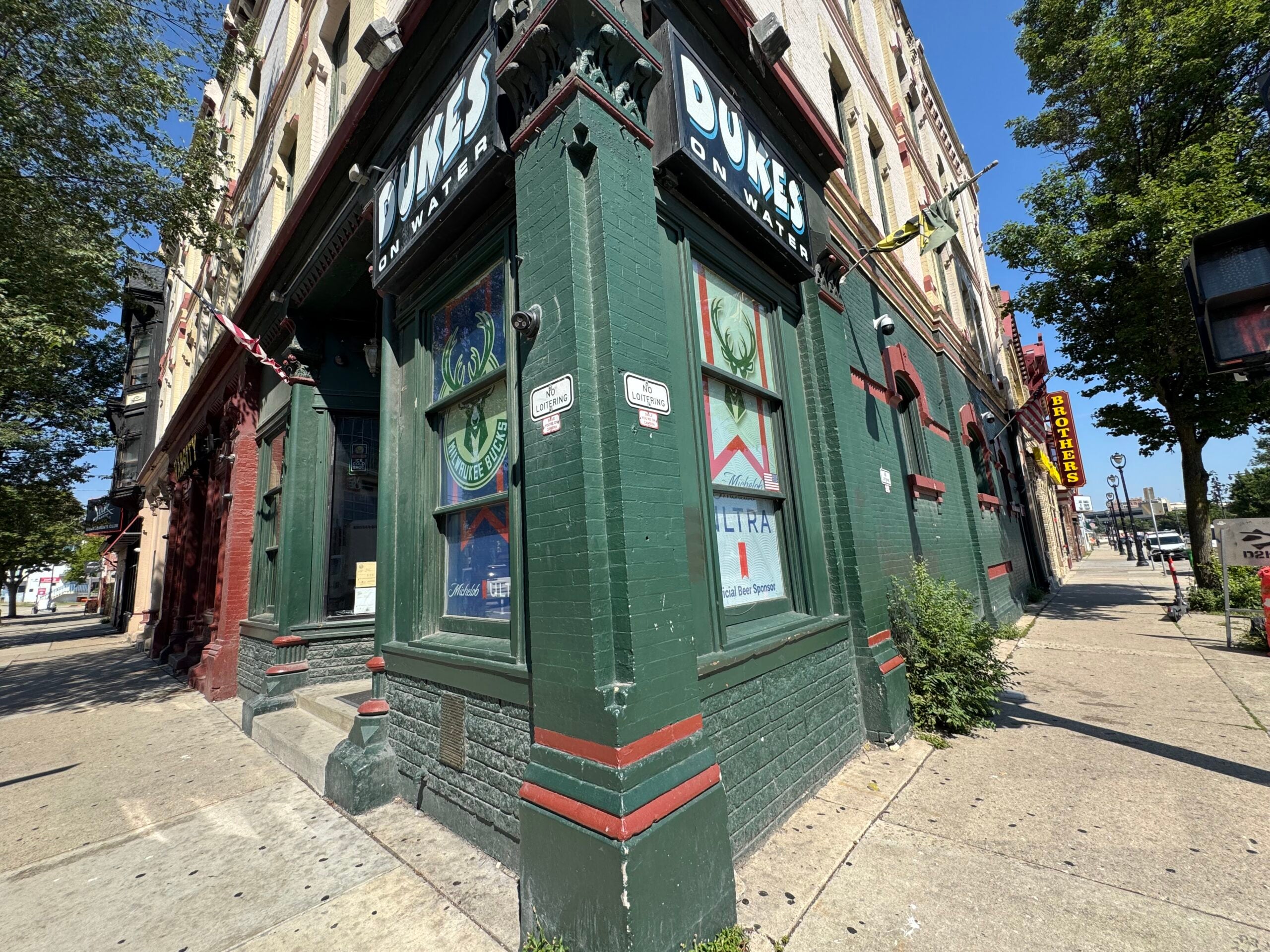 Corner view of a green and red brick building with “DUNES” signs above the windows, posters on the glass, and trees lining the sidewalk on a sunny day.