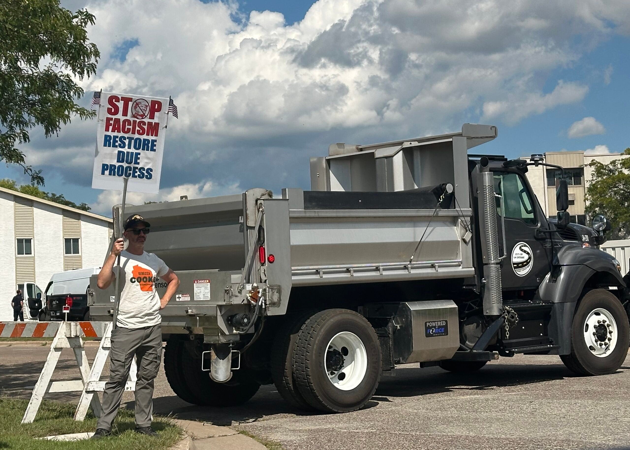 A person stands next to a dump truck holding a sign that reads, STOP FASCISM RESTORE DUE PROCESS on a sunny day with clouds in the sky.