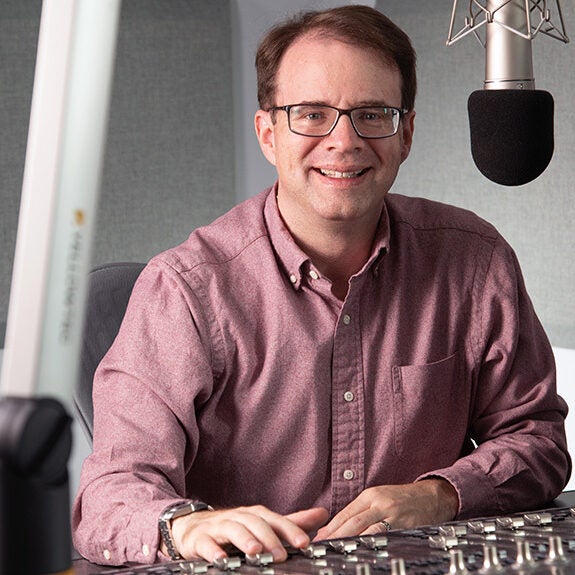 A man wearing glasses and a maroon shirt sits at a radio station control panel, smiling at the camera, with a microphone suspended above him.