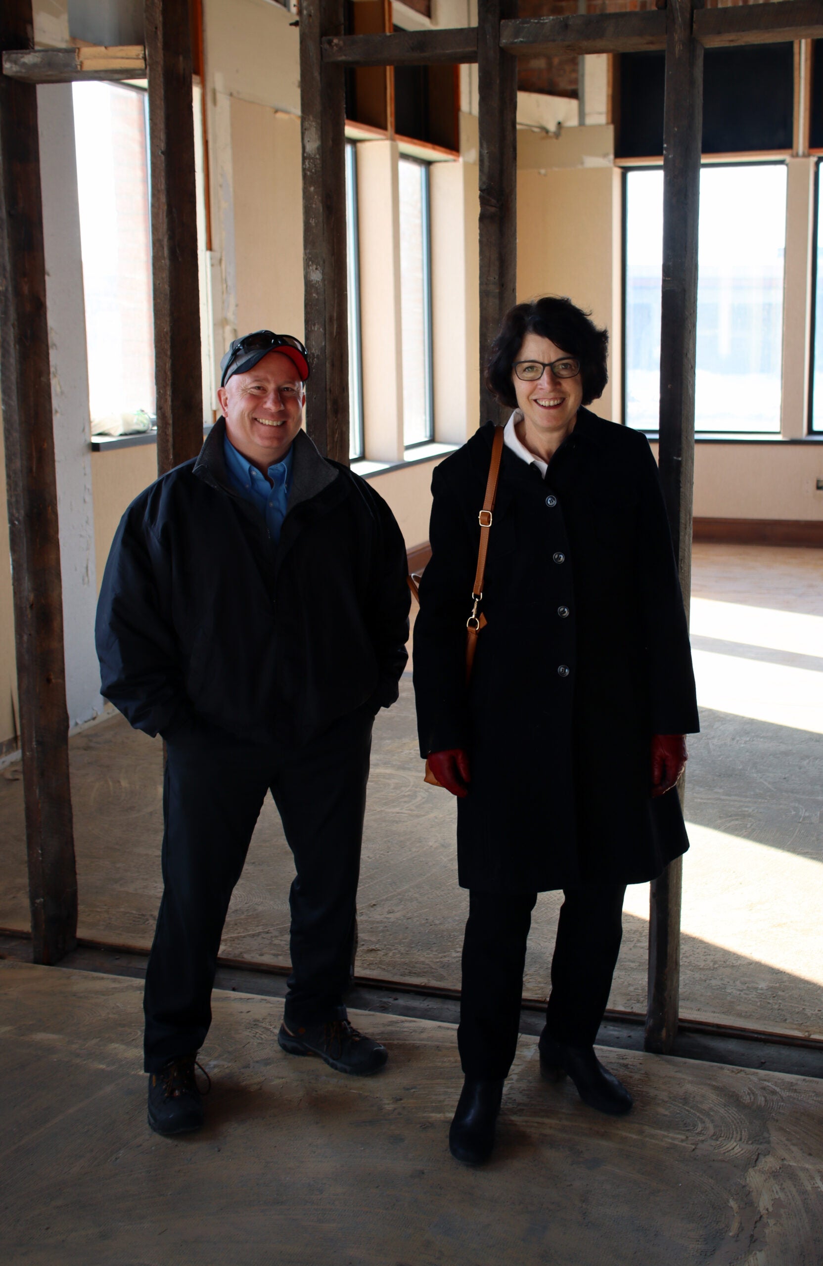 Two adults wearing coats stand and smile in a partially constructed or renovated room with exposed wooden beams and large windows.