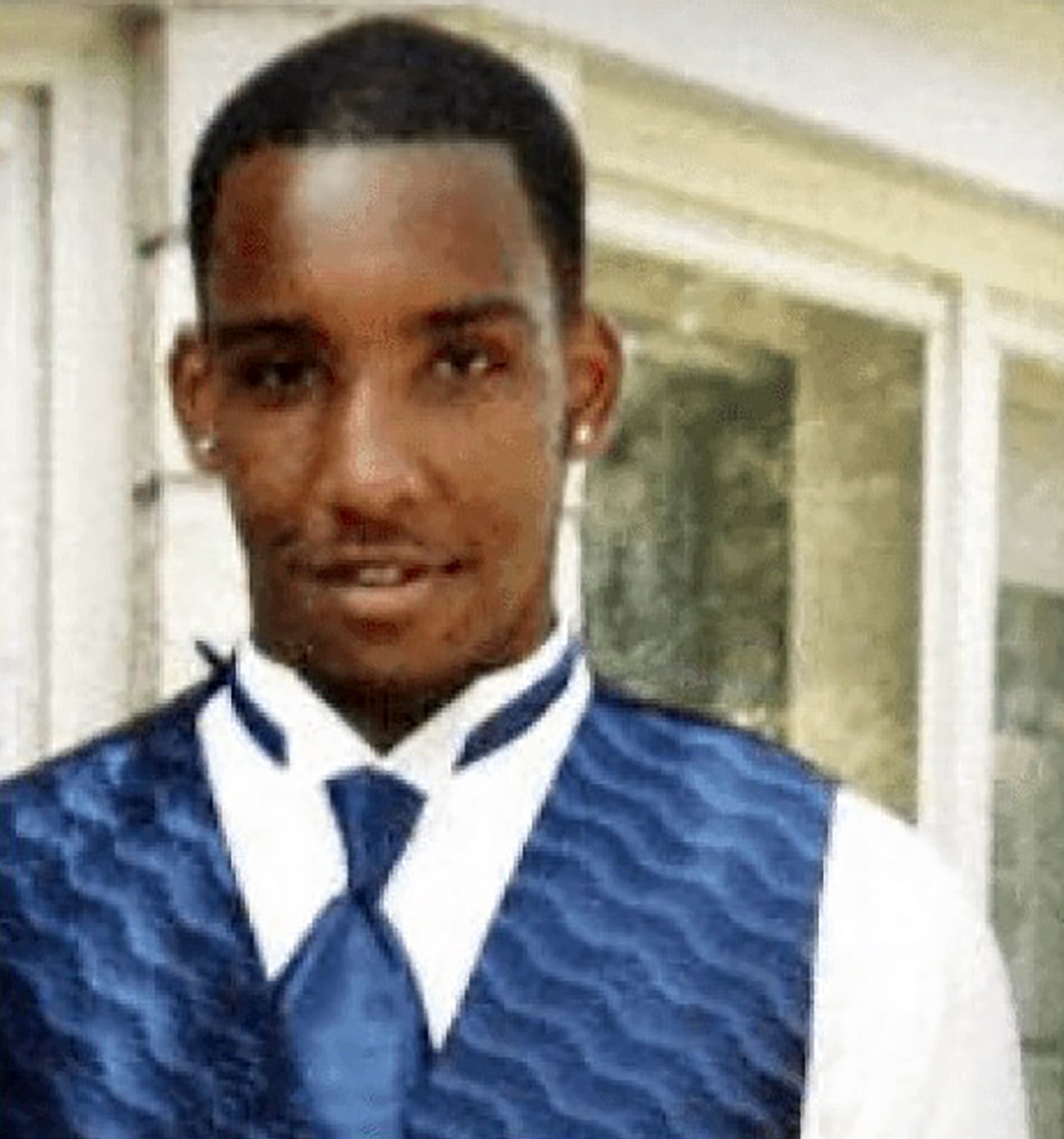 A man wearing a blue vest and tie over a white shirt stands indoors in front of a window, looking at the camera.