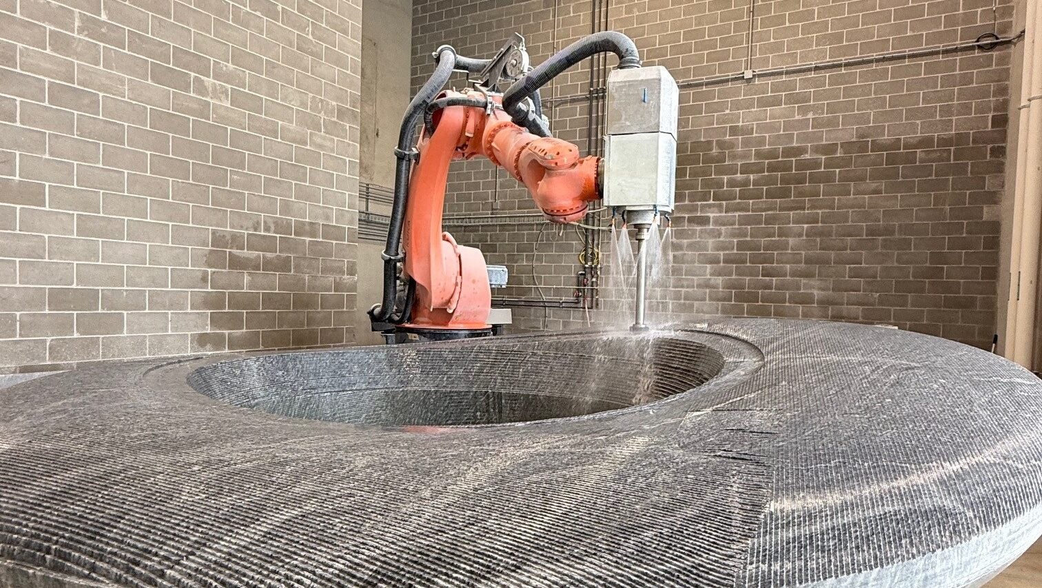 An industrial robotic arm carves a large circular stone slab in a workshop with brick walls and high ceilings.