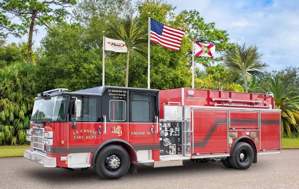 A red La Crosse Fire Dept. fire truck is parked on a road, with three flags flying behind it and trees in the background.