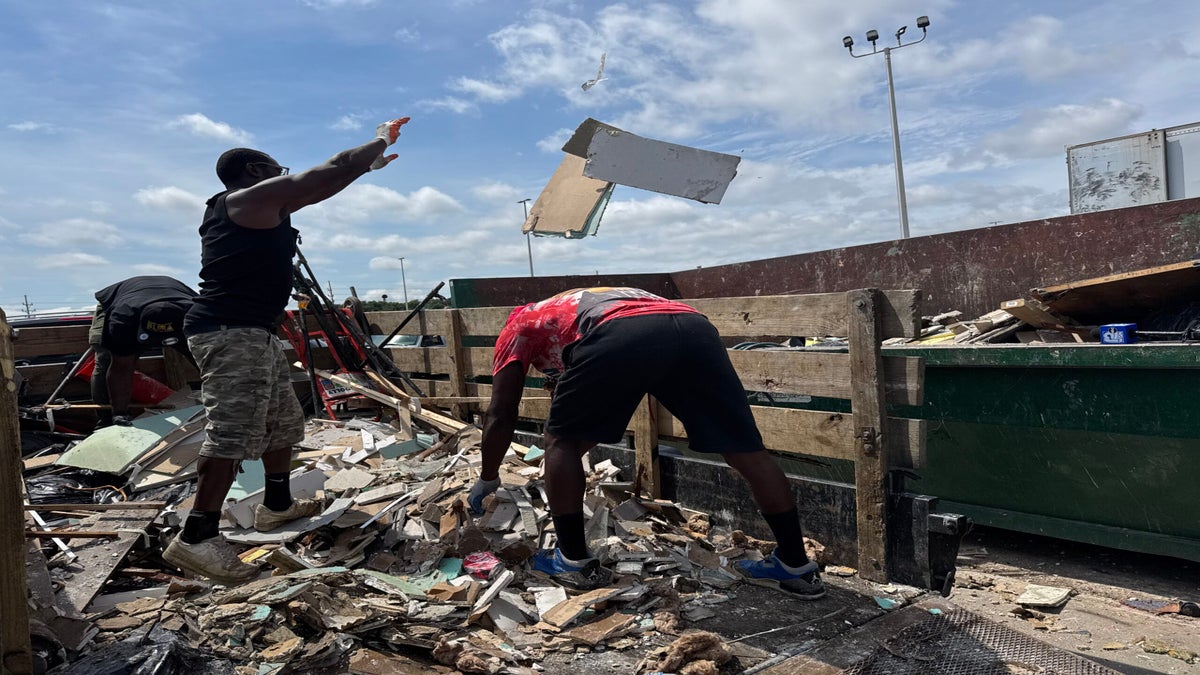 Two people unload and throw construction debris into a large dumpster at a junkyard under a partly cloudy sky.