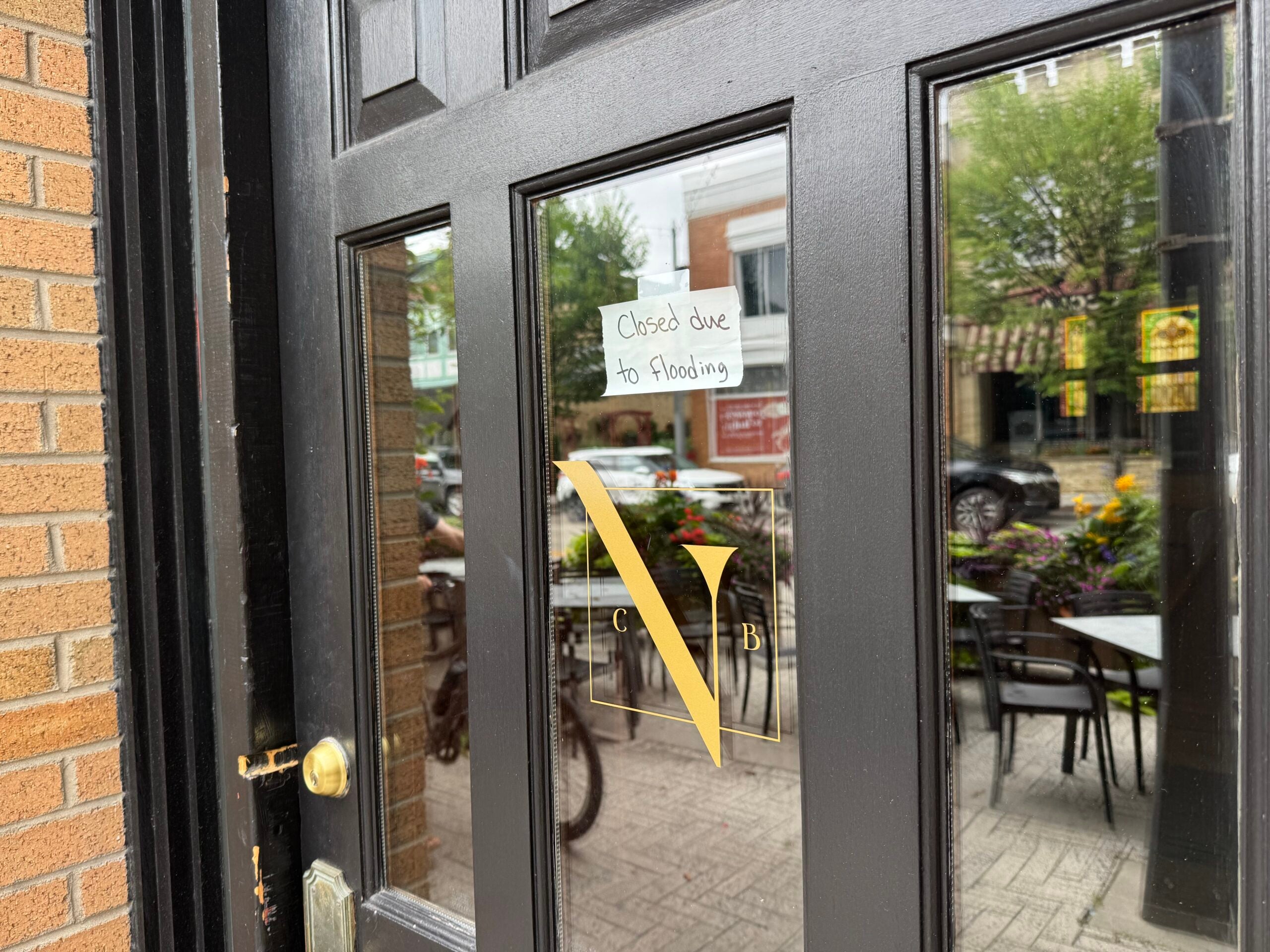 A handwritten sign on a glass door reads Closed due to flooding. Outdoor tables and chairs are visible through the door.
