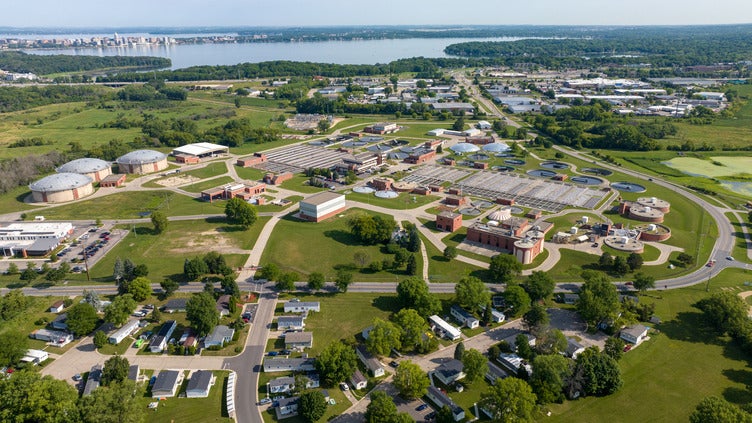 Aerial view of a water treatment facility with large circular tanks, surrounded by greenery, roads, and a nearby residential area, with a lake and city in the background.