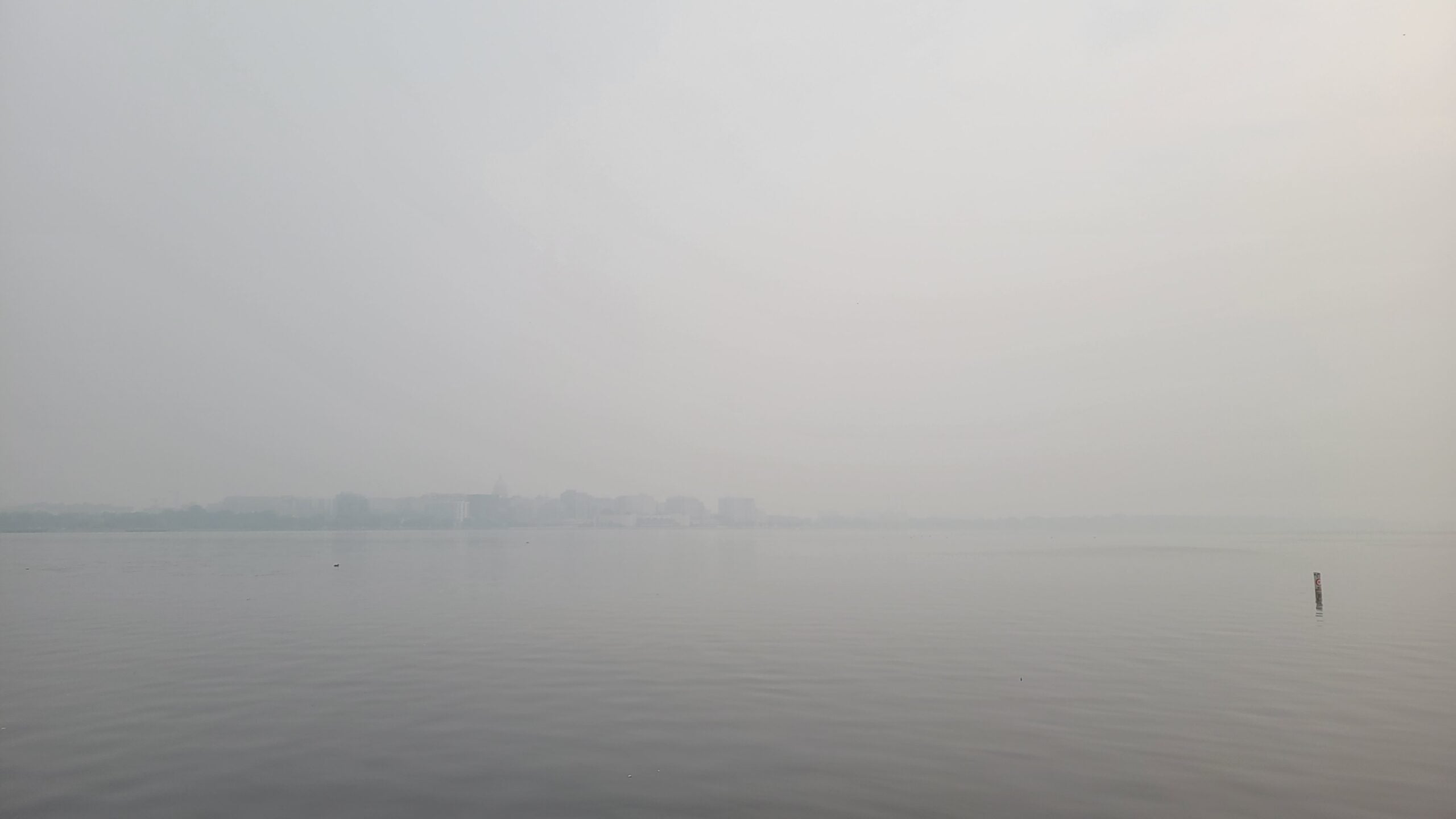 A hazy sky over a calm body of water, with distant buildings barely visible through the thick fog or smog.