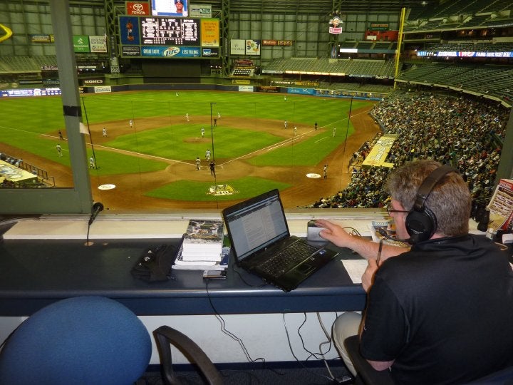 A person wearing headphones sits in a broadcast booth overlooking a baseball game, using a laptop and microphone, with the field and spectators visible through the window.