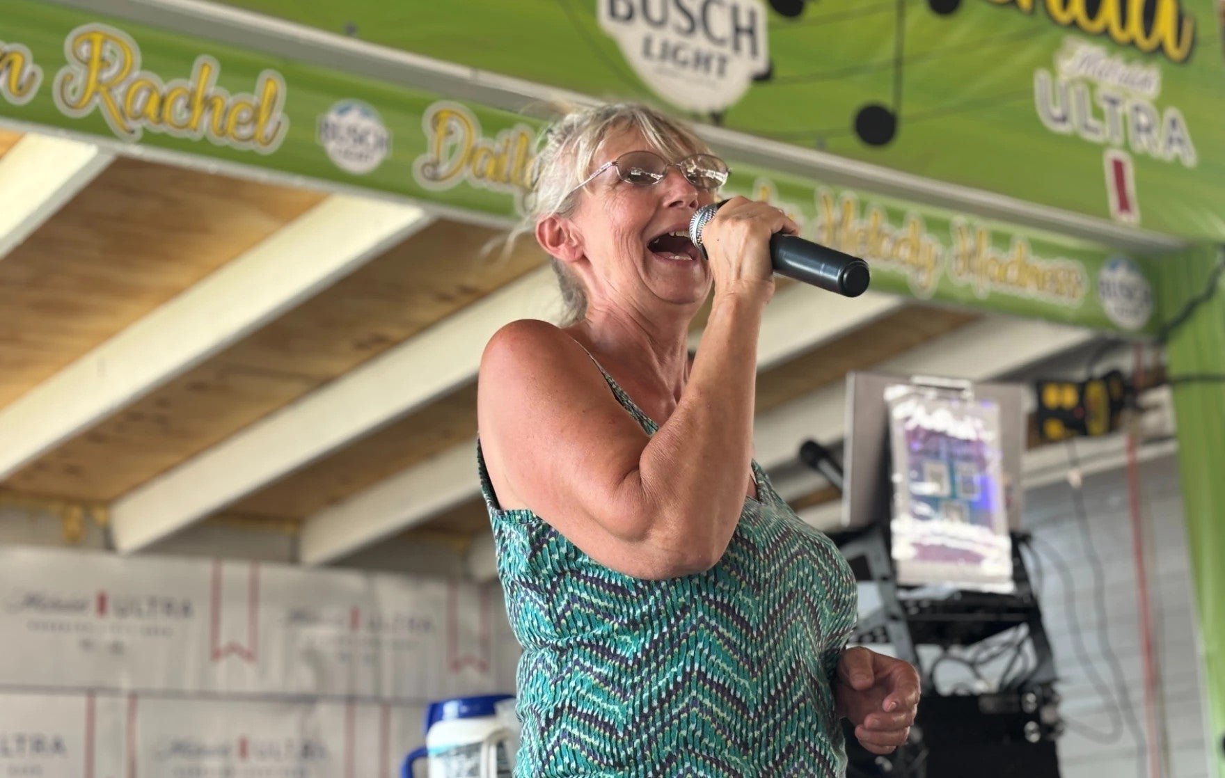 An older woman in glasses and a patterned dress sings into a microphone on an indoor-outdoor stage decorated with beer advertisements.