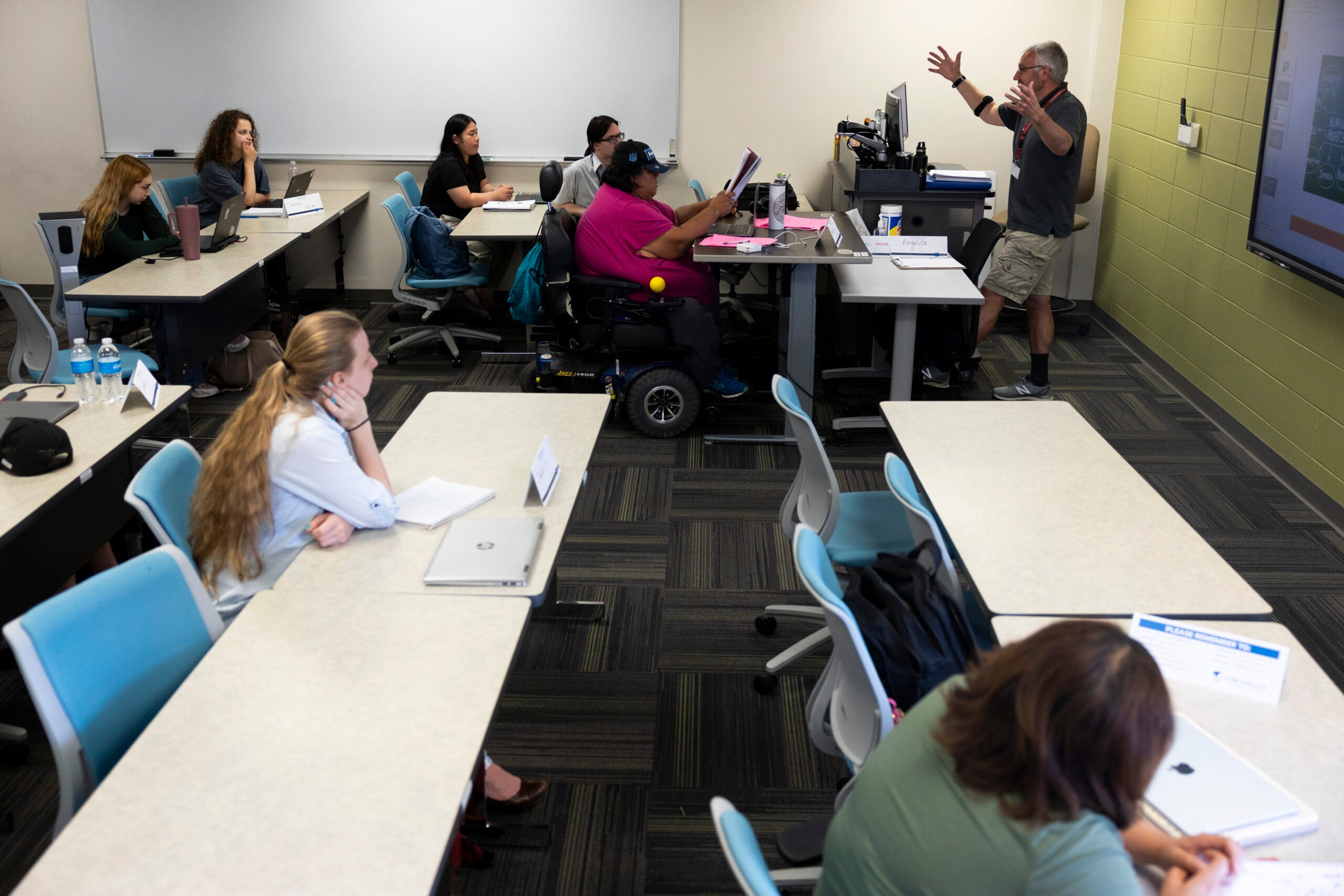 A classroom scene with a teacher speaking at the front and several students seated at desks, some with laptops, listening and taking notes.