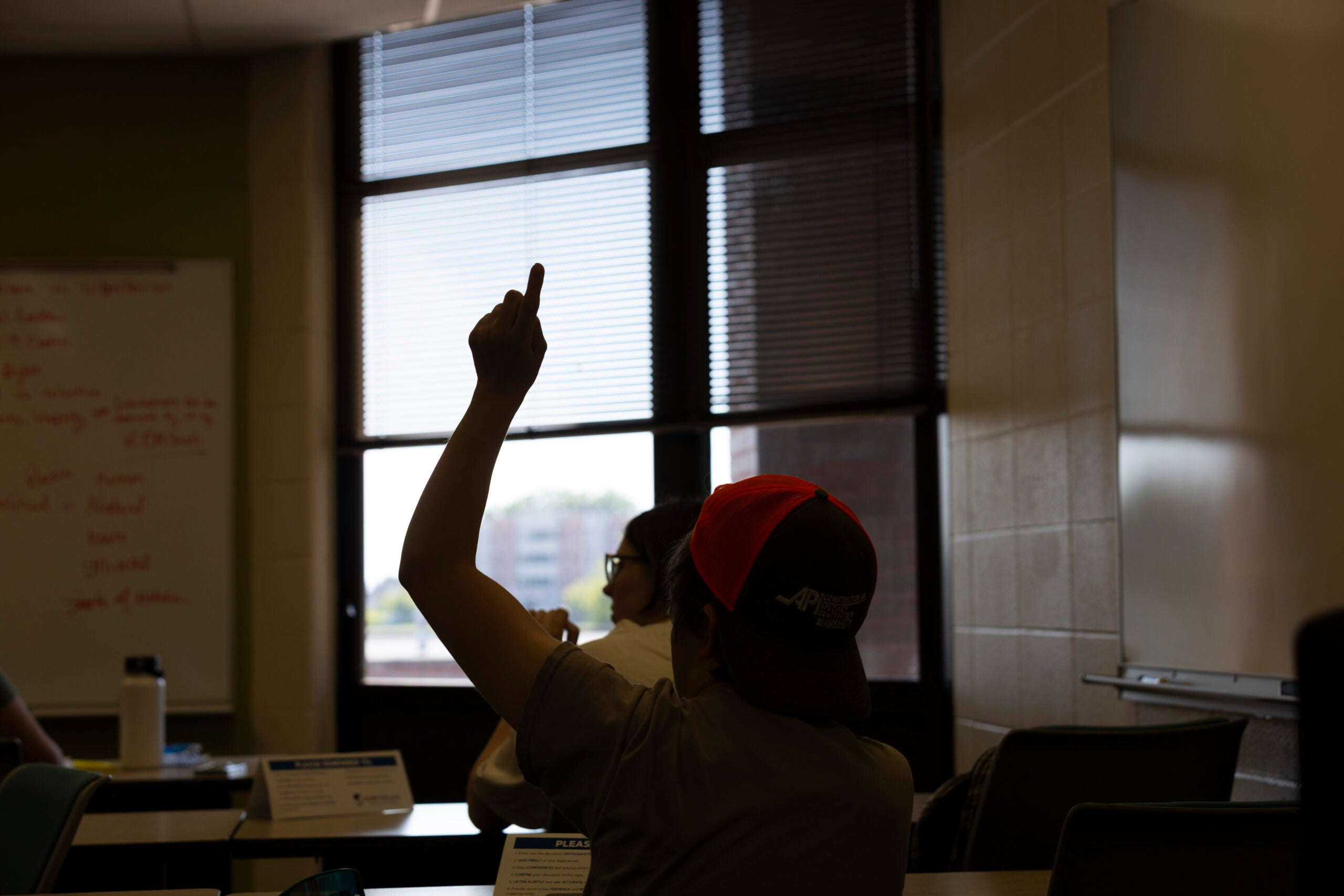 A person wearing a red cap raises their hand in a classroom with large windows and a whiteboard in the background.