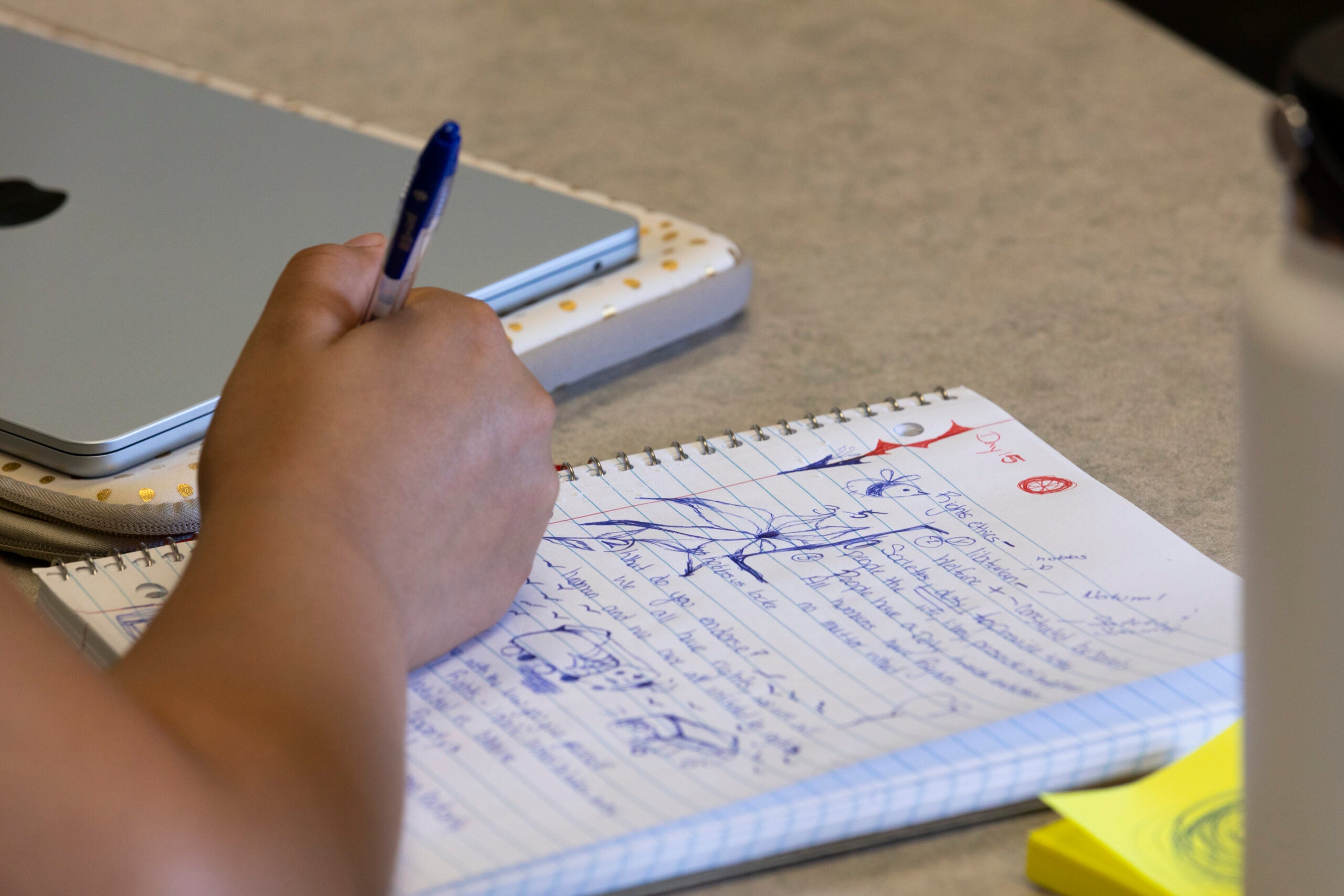 A person writes notes with a blue pen in a spiral notebook beside a closed laptop and some sticky notes on a desk.