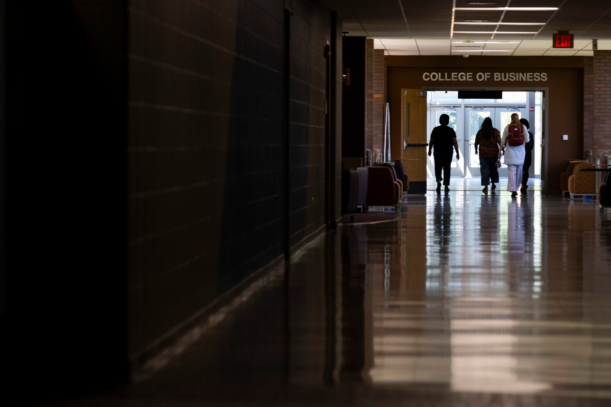 Four people walk down a hallway toward a door labeled College of Business in a building with shiny floors and brick walls.