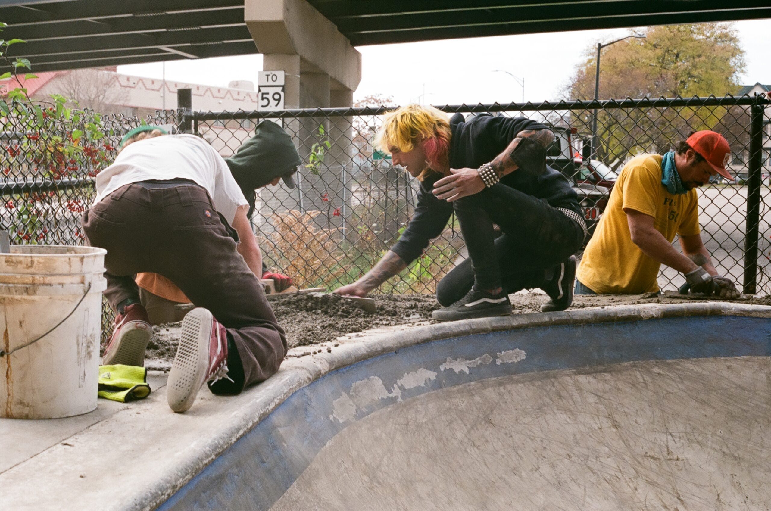 Three people patch concrete at the edge of a skate bowl under a bridge, with tools, a bucket, and a chain-link fence in the background.