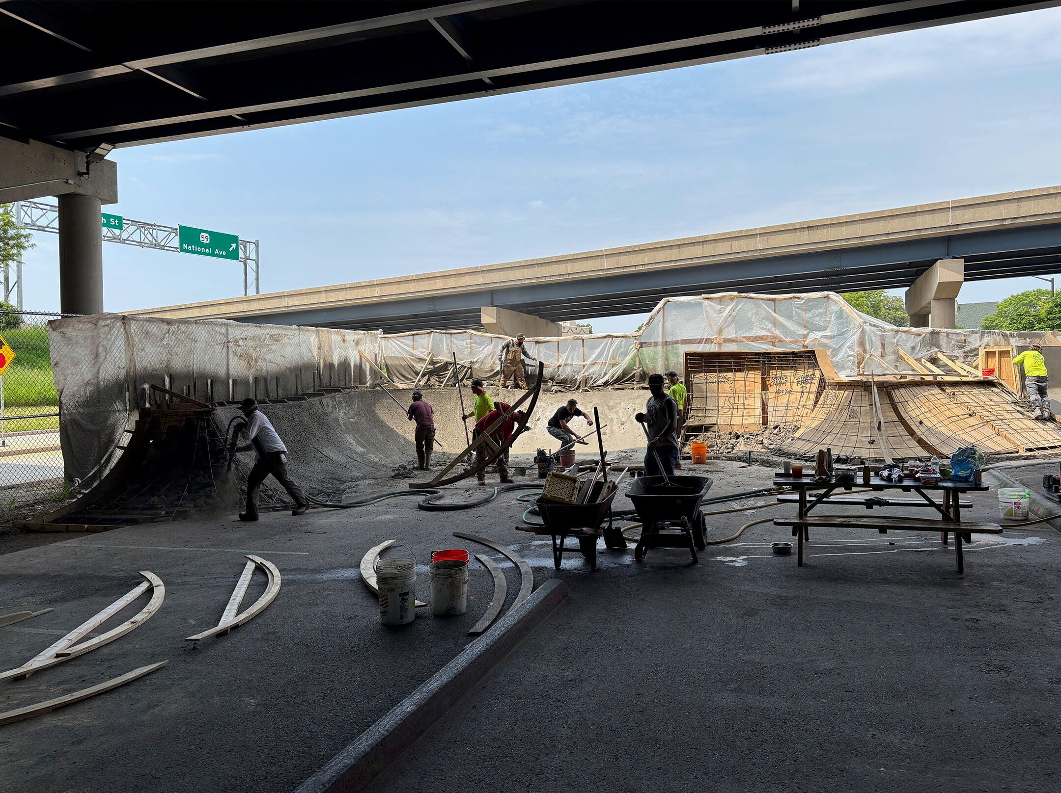 Construction workers building and shaping a large concrete skatepark under a highway overpass, with tools and materials scattered nearby.