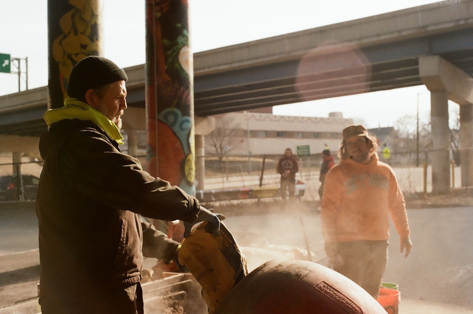 Two people work outdoors near a highway overpass, one pouring material into a drum as dust rises; painted pillars and urban structures are visible in the background.
