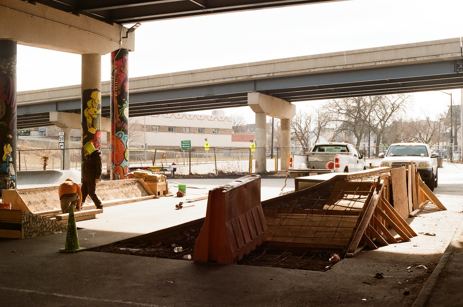 A construction zone under an overpass with traffic barriers, fencing, and debris; two parked trucks and a person walking nearby.