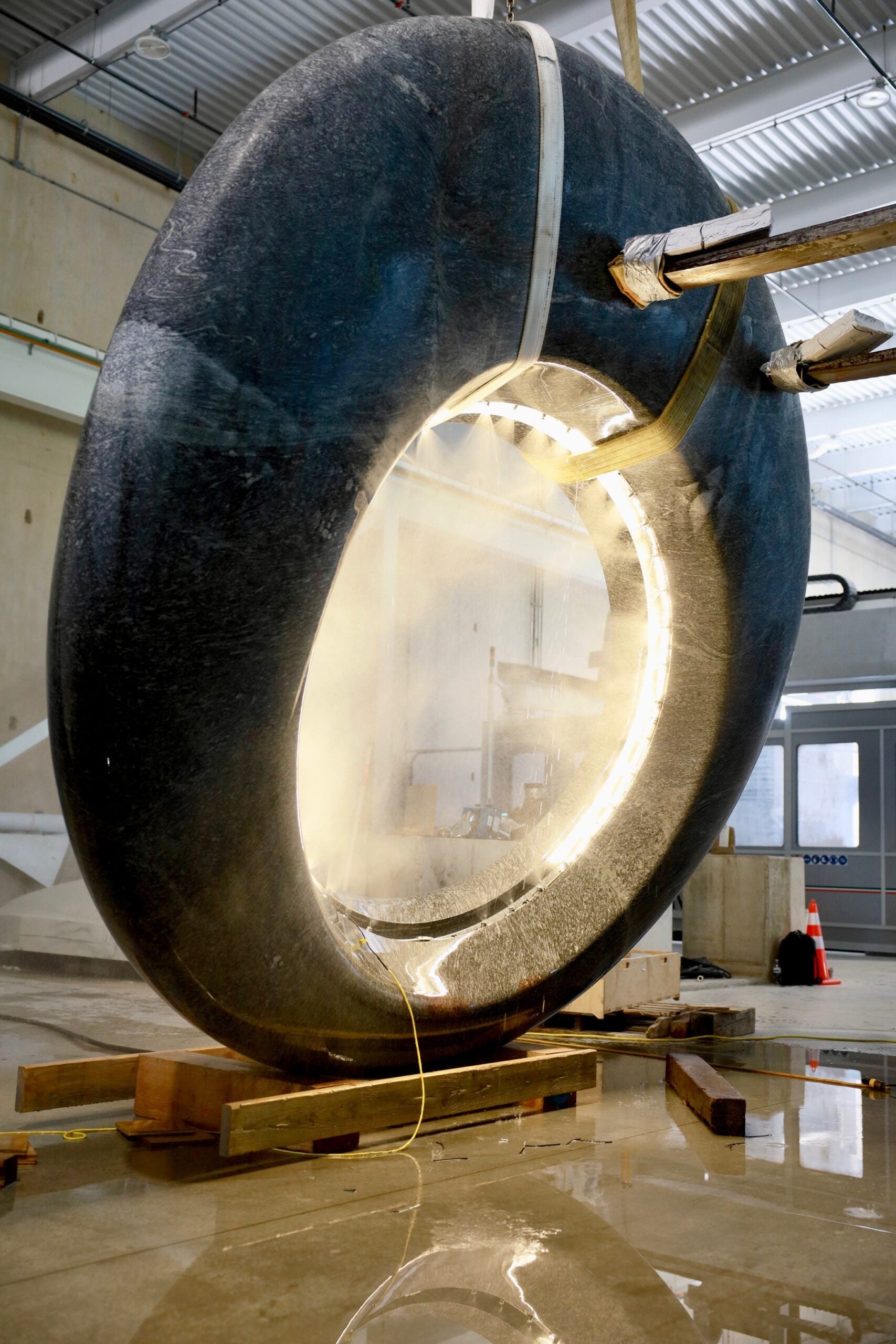 A large circular structure is suspended by metal supports, with mist or steam emanating from its inner surface, inside an industrial facility.